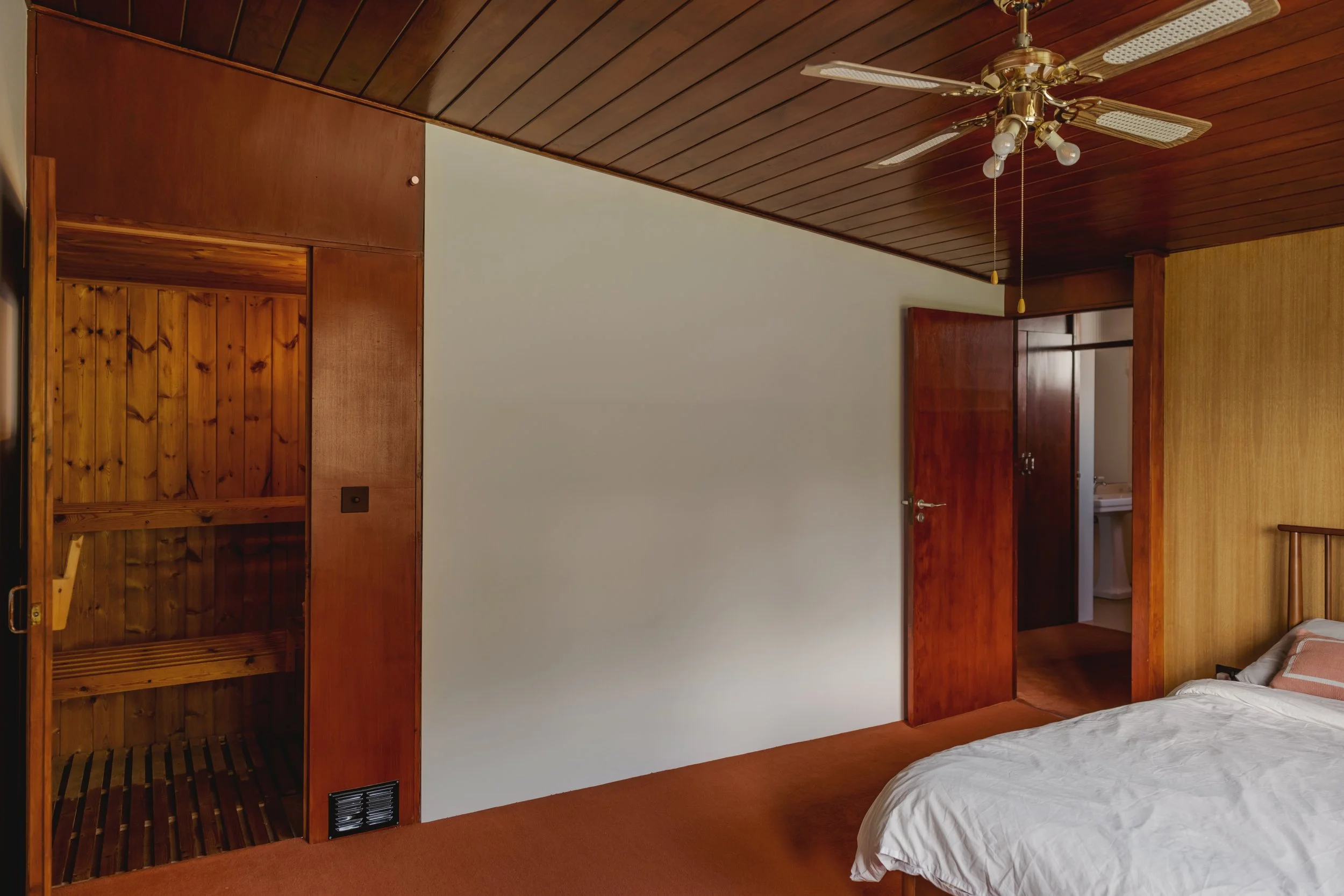 A bedroom with wood-paneled ceiling and walls, a brass ceiling fan, a white bed with a pillow, an open closet with shelves, and a door leading to a small bathroom or sink area.