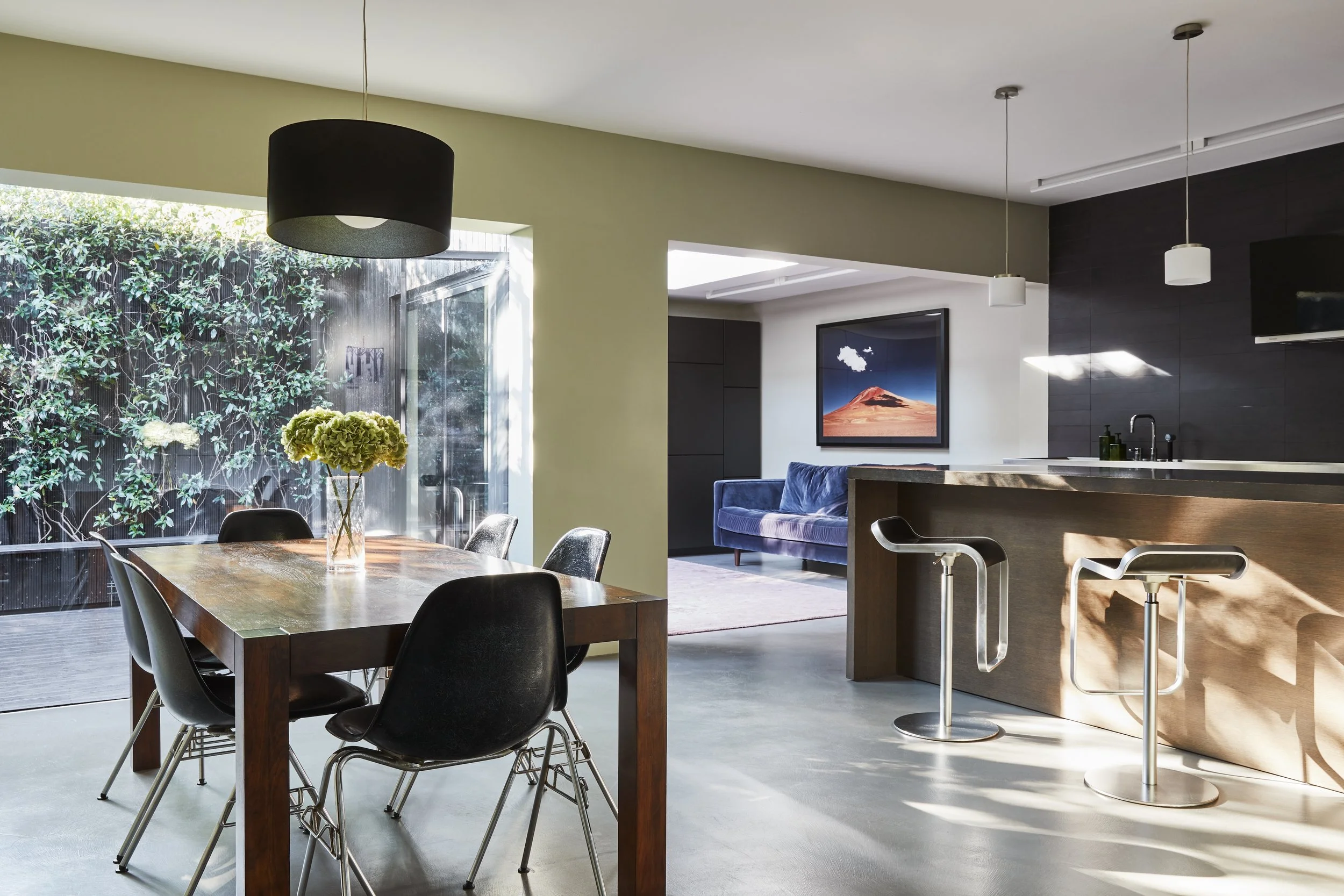 Modern open-plan kitchen and dining area with wooden dining table, black chairs, glass vase with flowers, bar stools, dark cabinetry, artwork of a mountain, and large windows letting in natural light.
