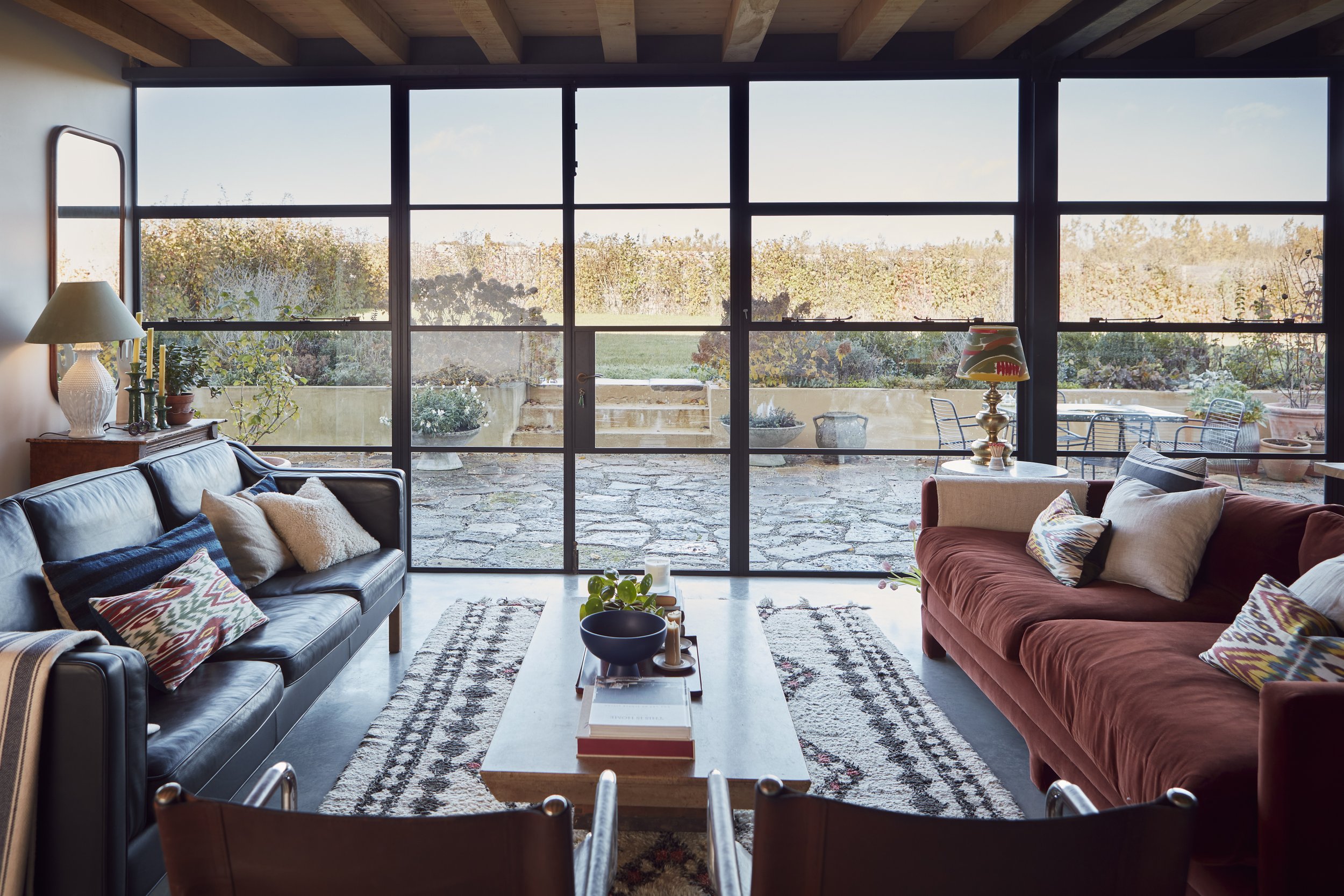 Living room with large window view of garden, two sofas with cushions, coffee table with books, lamp, and side tables with plants and candles.