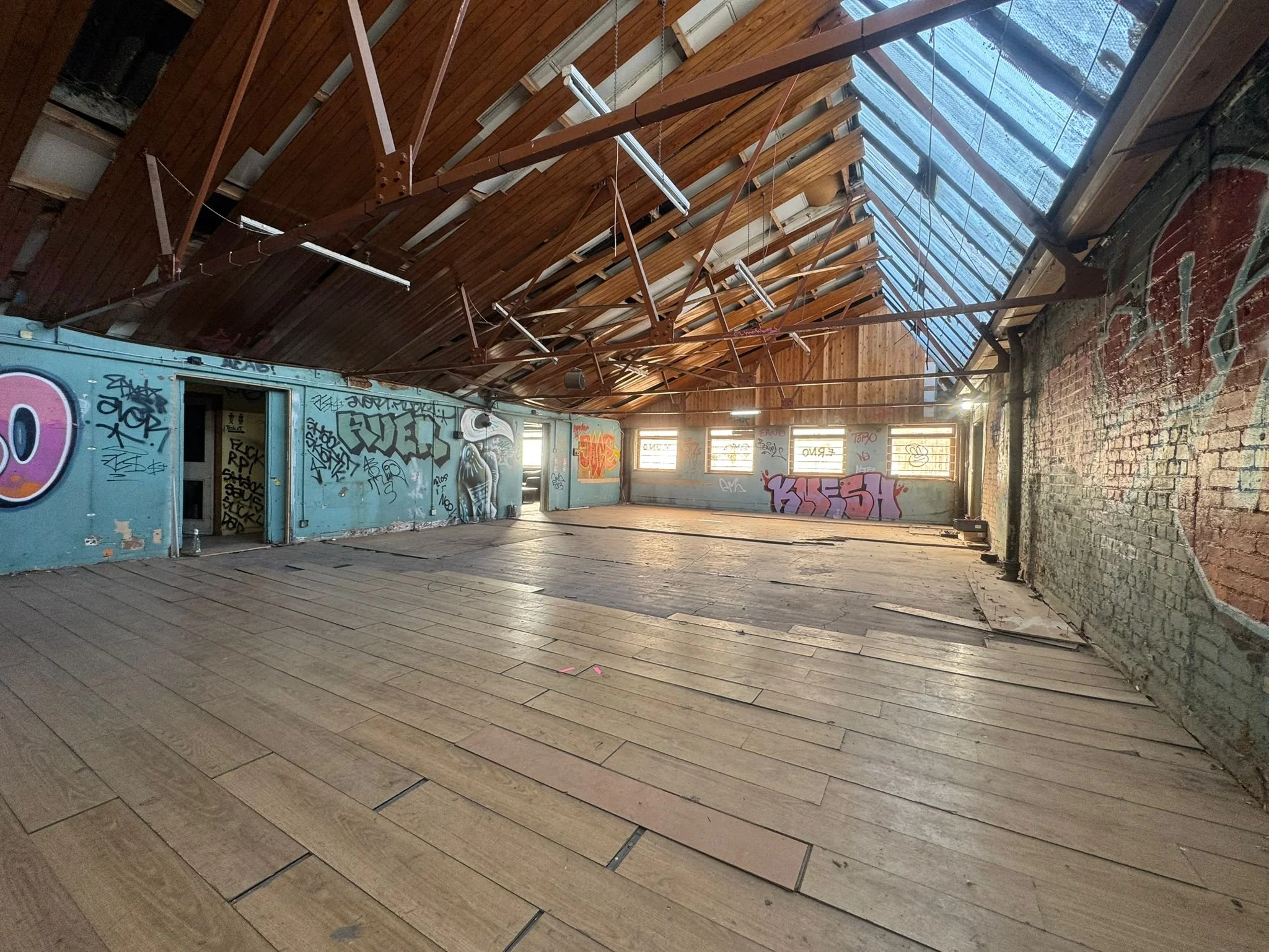 An interior space with wooden flooring, graffiti-covered walls, graffiti, and a roof with exposed wooden beams and glass panels. The space appears abandoned and is lit by natural light.