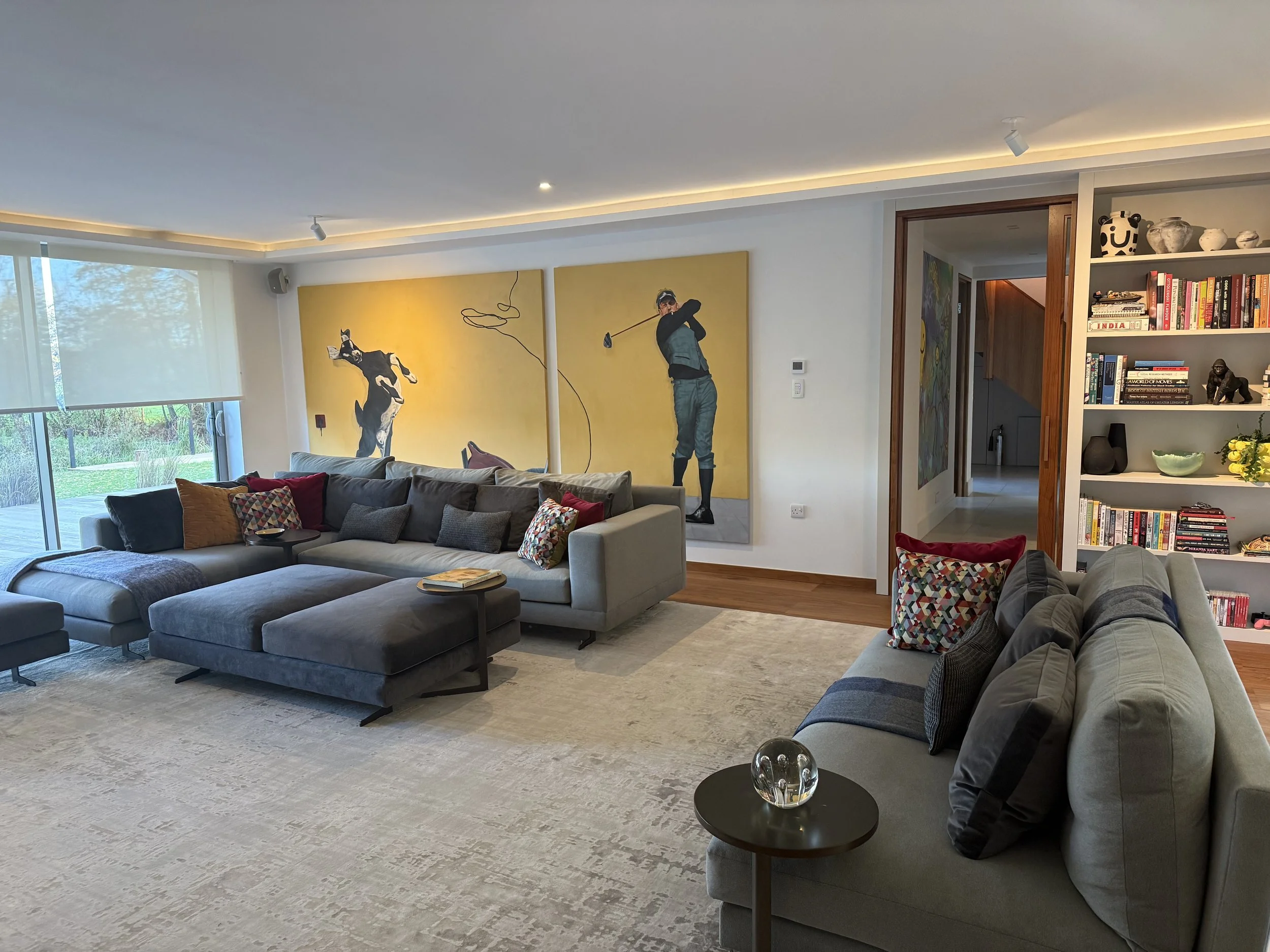 A spacious living room with a large gray sectional sofa, colorful throw pillows, and a beige armchair. Large window with blinds showing outdoor greenery. Modern art paintings on the wall. Open doorway leading to a hallway. Shelf with books and decora