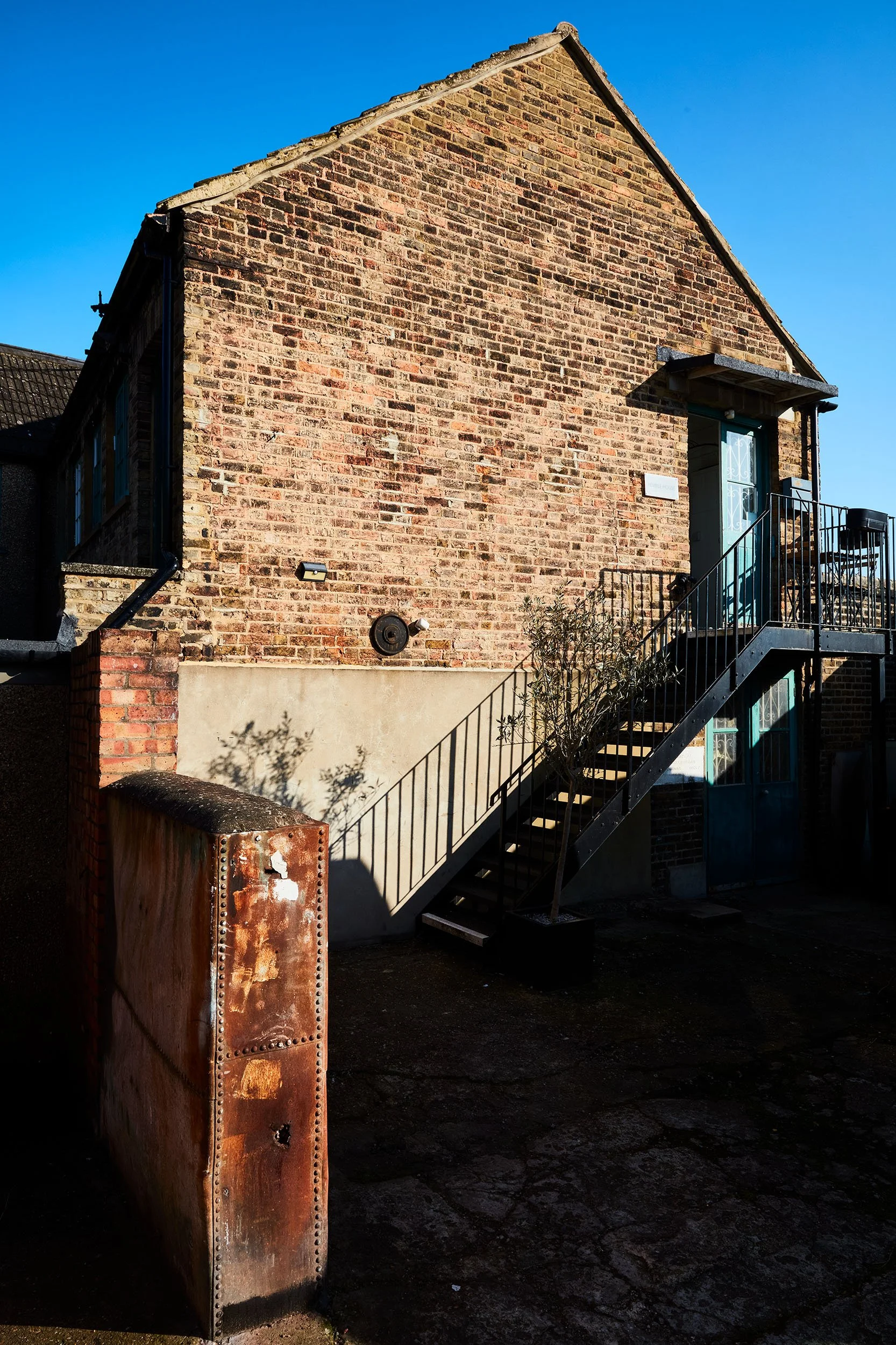 A brick building with a staircase leading to an upper door, shadows cast by a nearby tree, and a rusty metal box in the foreground under a clear blue sky.