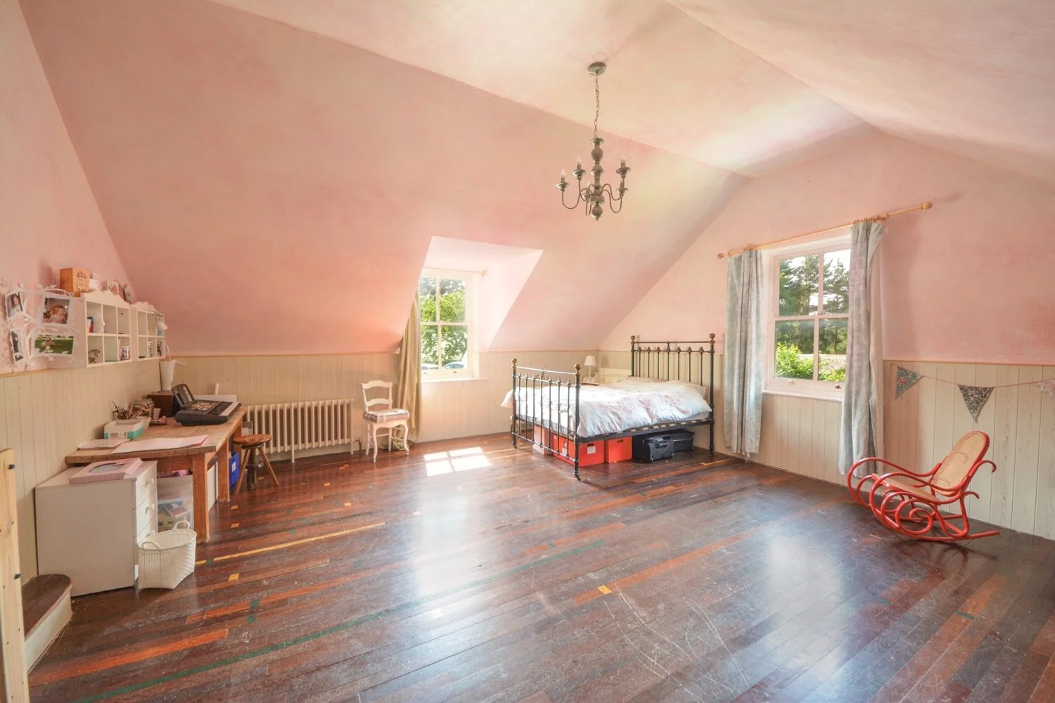 A spacious attic bedroom with pink painted sloped ceiling, hardwood floor, two windows with curtains, an antique metal bed with white bedding, a red wooden rocking chair, a small desk with a printer, and various decorations including a chandelier and