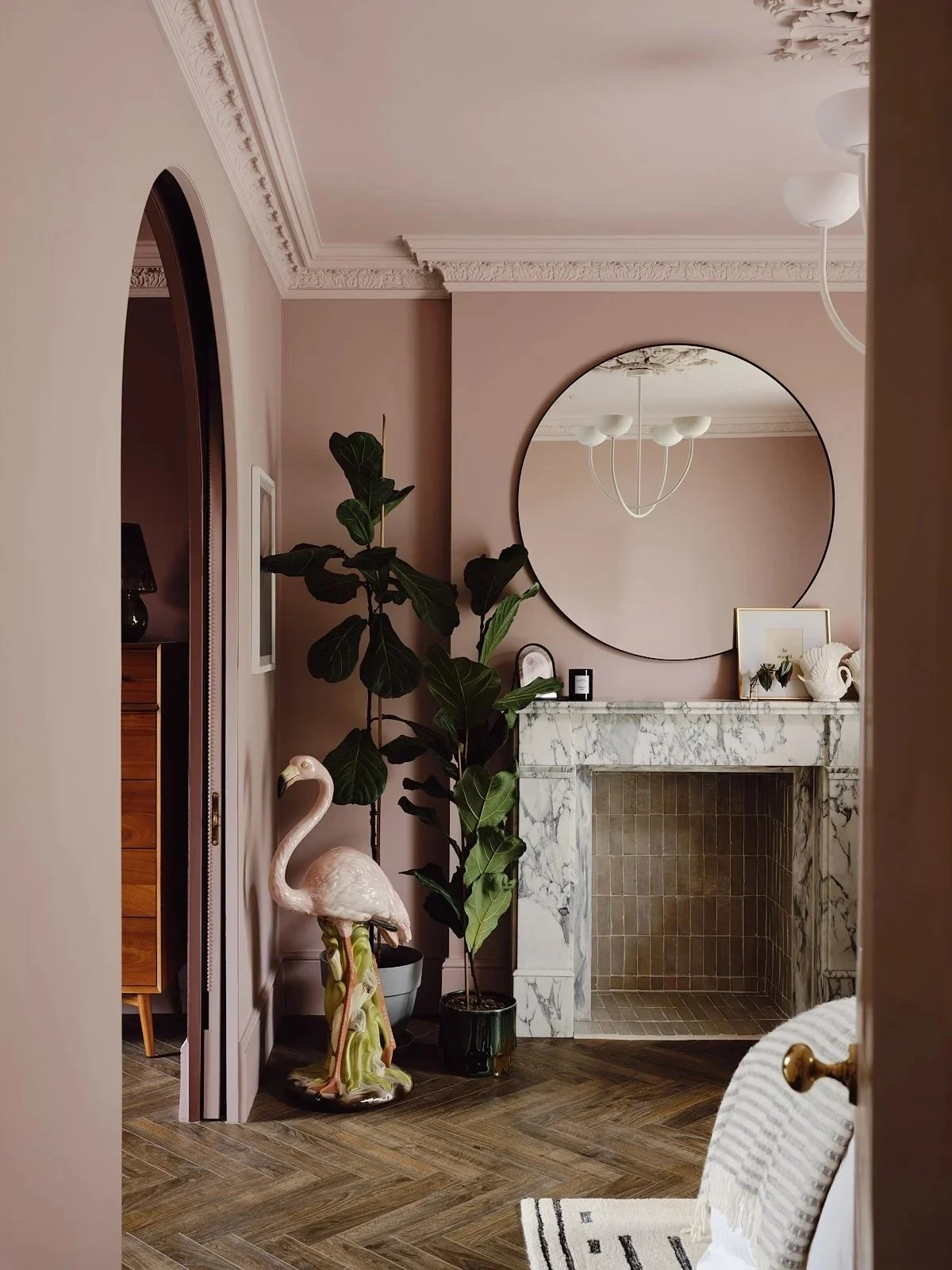 Living room corner with pink walls, decorative white molding, a round mirror above a fireplace with a marble mantel, a large green houseplant next to a flamingo statue, and part of a striped rug and a wooden cabinet visible.