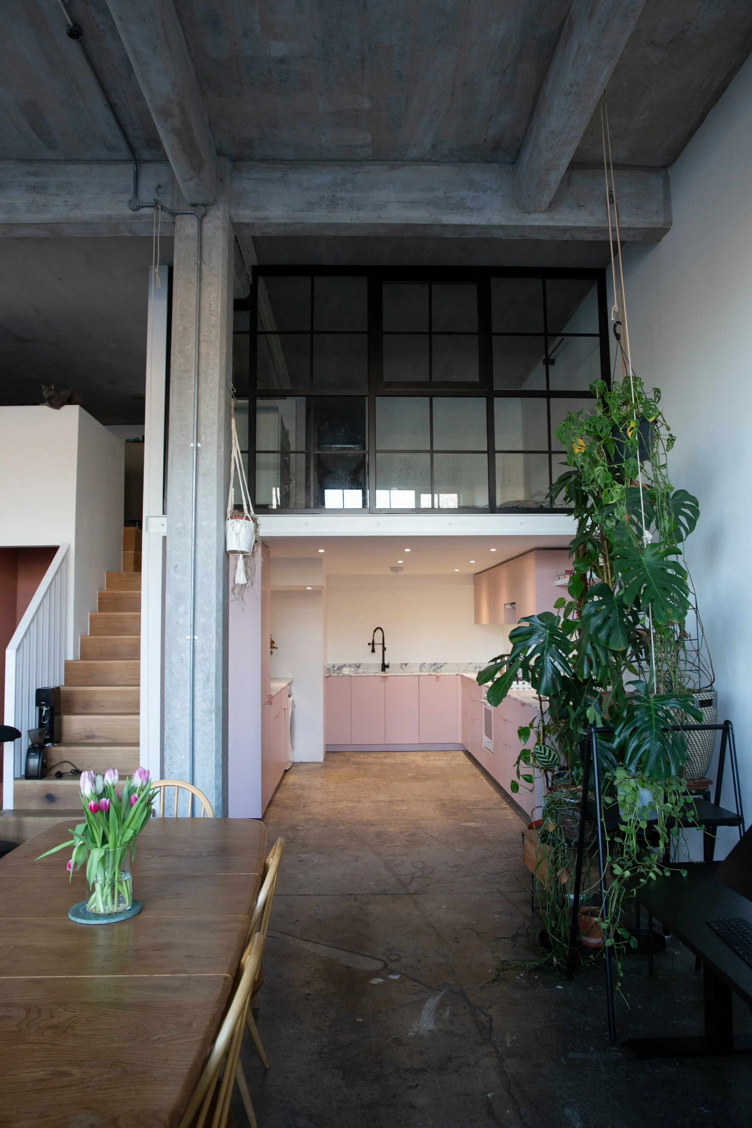 Interior view of a modern, industrial-style loft apartment with an open kitchen, pink cabinets, and a balcony with black framing. There is a wooden dining table with a vase of pink tulips, large indoor plants, and concrete flooring.