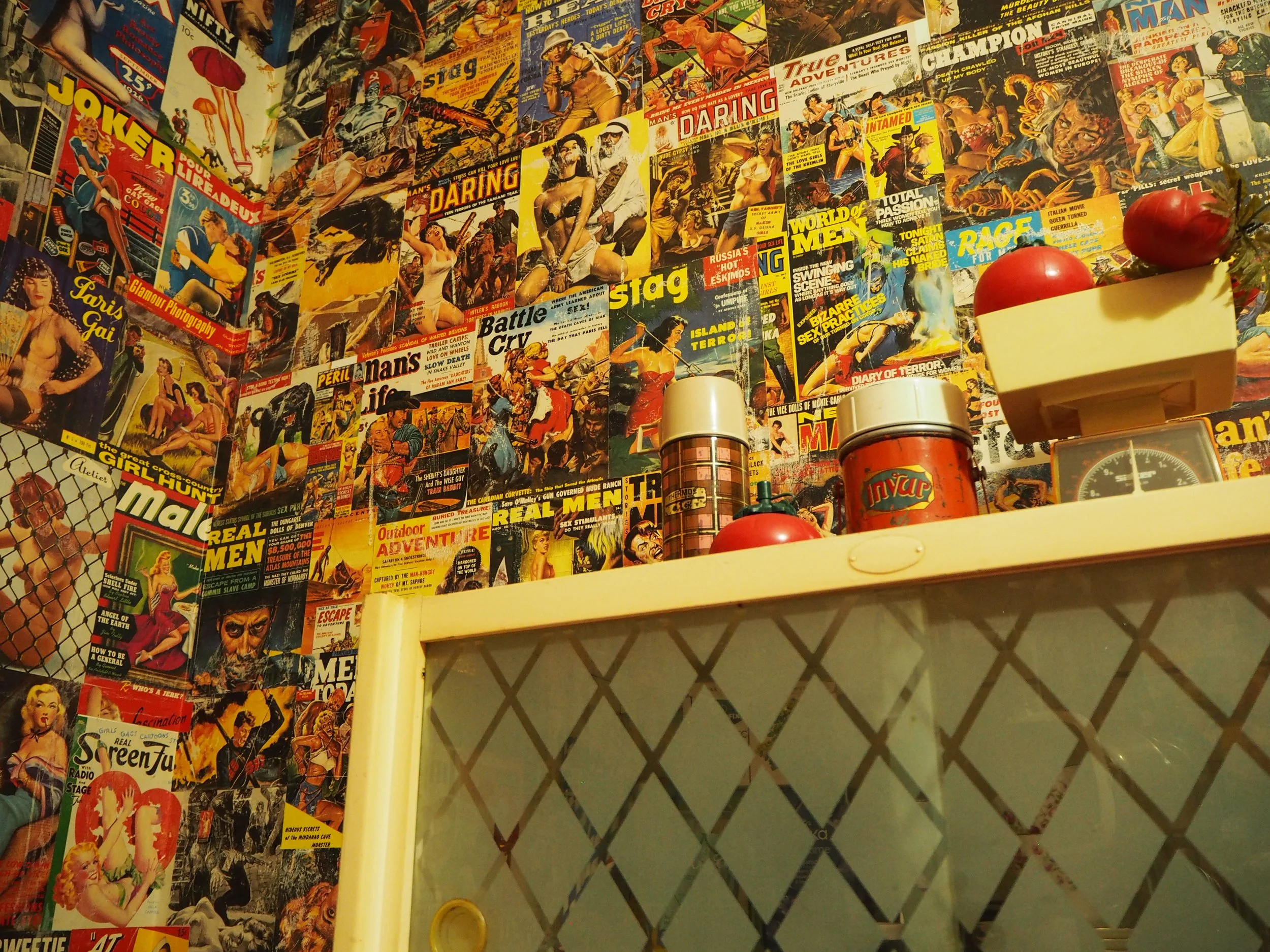 A wall covered with vintage magazine covers featuring women, adventure, and action-themed illustrations, with a small section of a yellow shelf holding cans, jars, and tomatoes, and a scale visible on the right side.