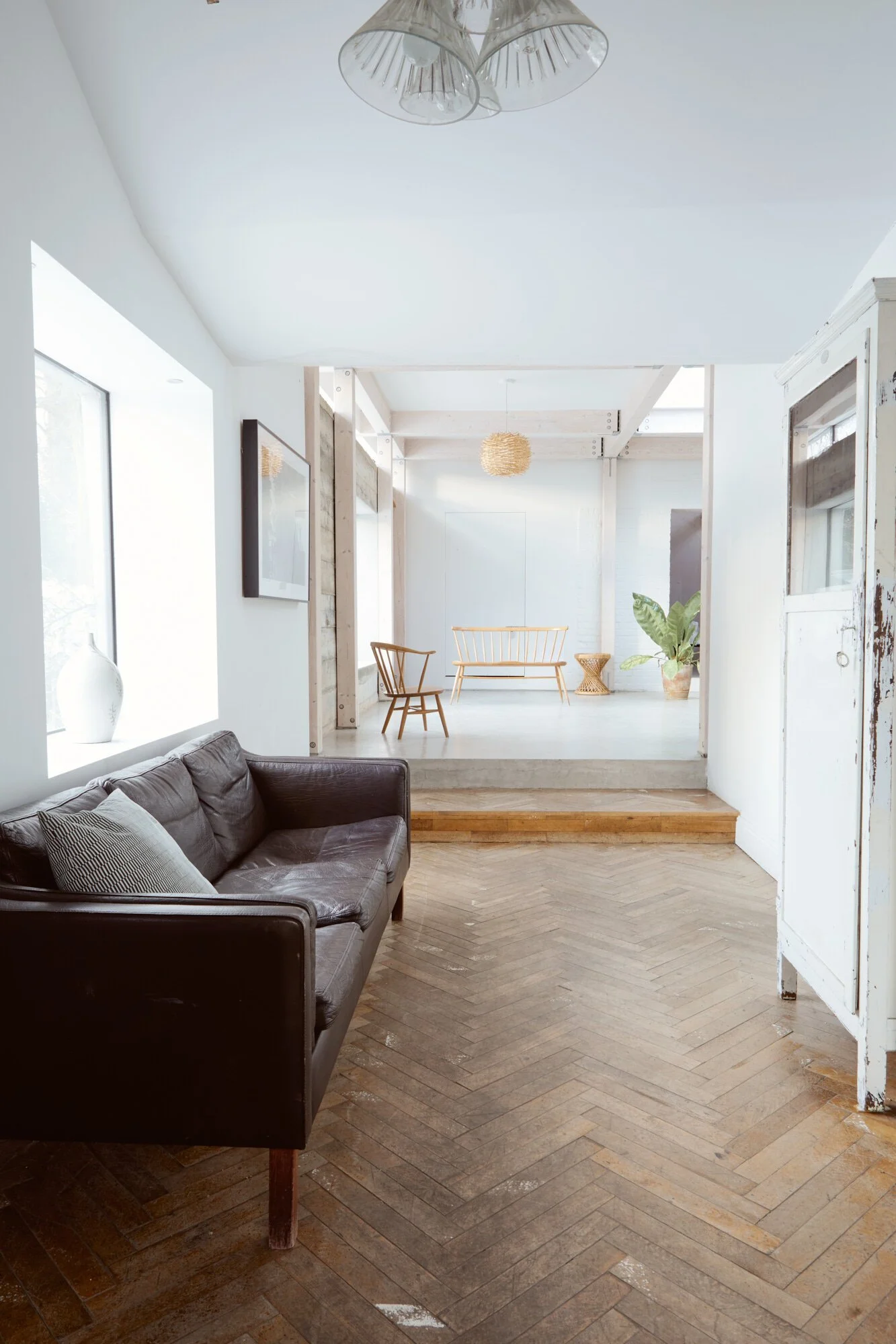 Living room with hardwood herringbone flooring, a dark leather sofa with a pillow, a window with natural light, and a work of art on the wall. In the background are a raised platform with light-colored flooring, various chairs, a plant, and a hanging