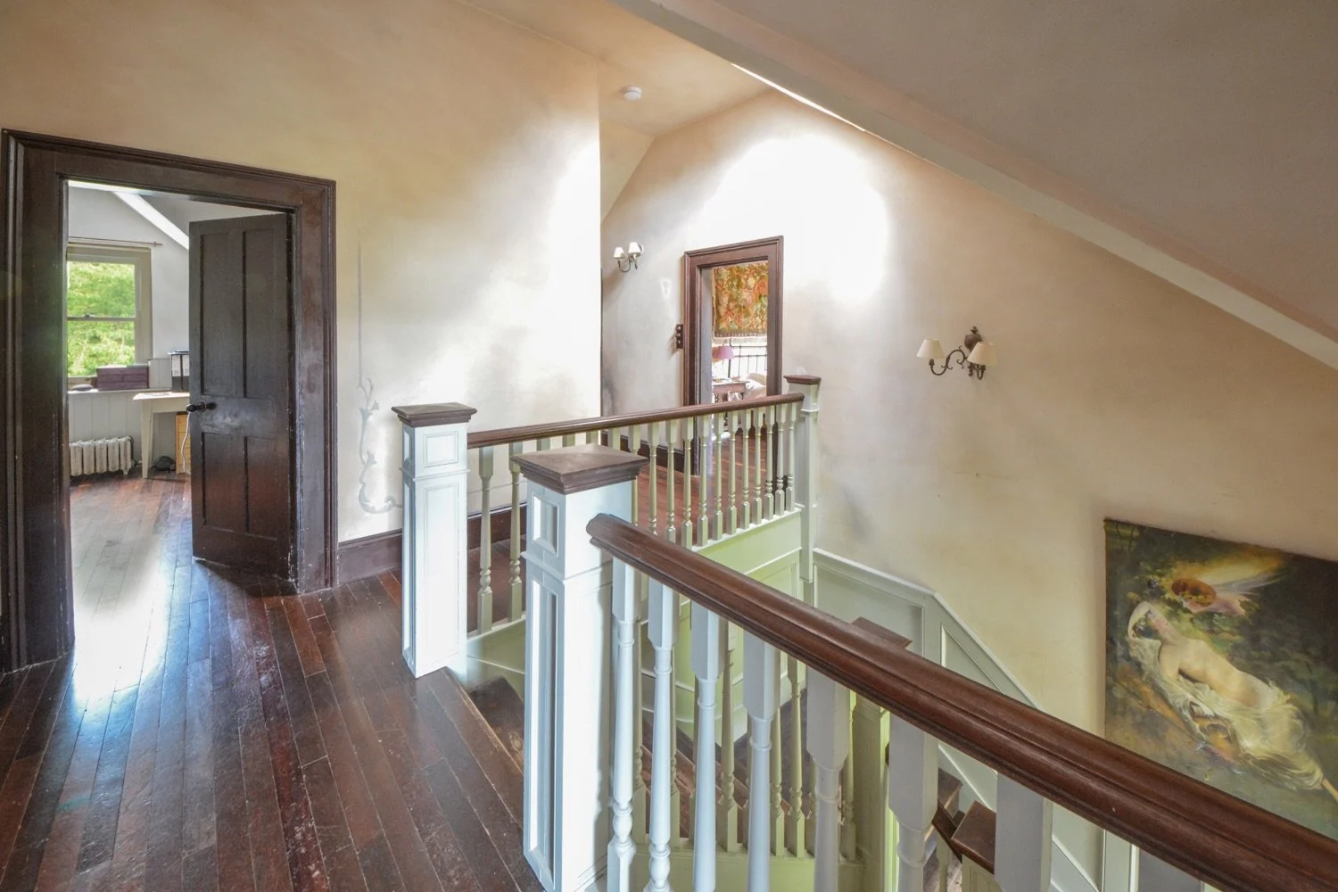 Photo of an interior staircase landing with dark wooden floors, white railing, and walls with artistic paintings. There are doors leading to various rooms, and natural light coming from a window in the background.