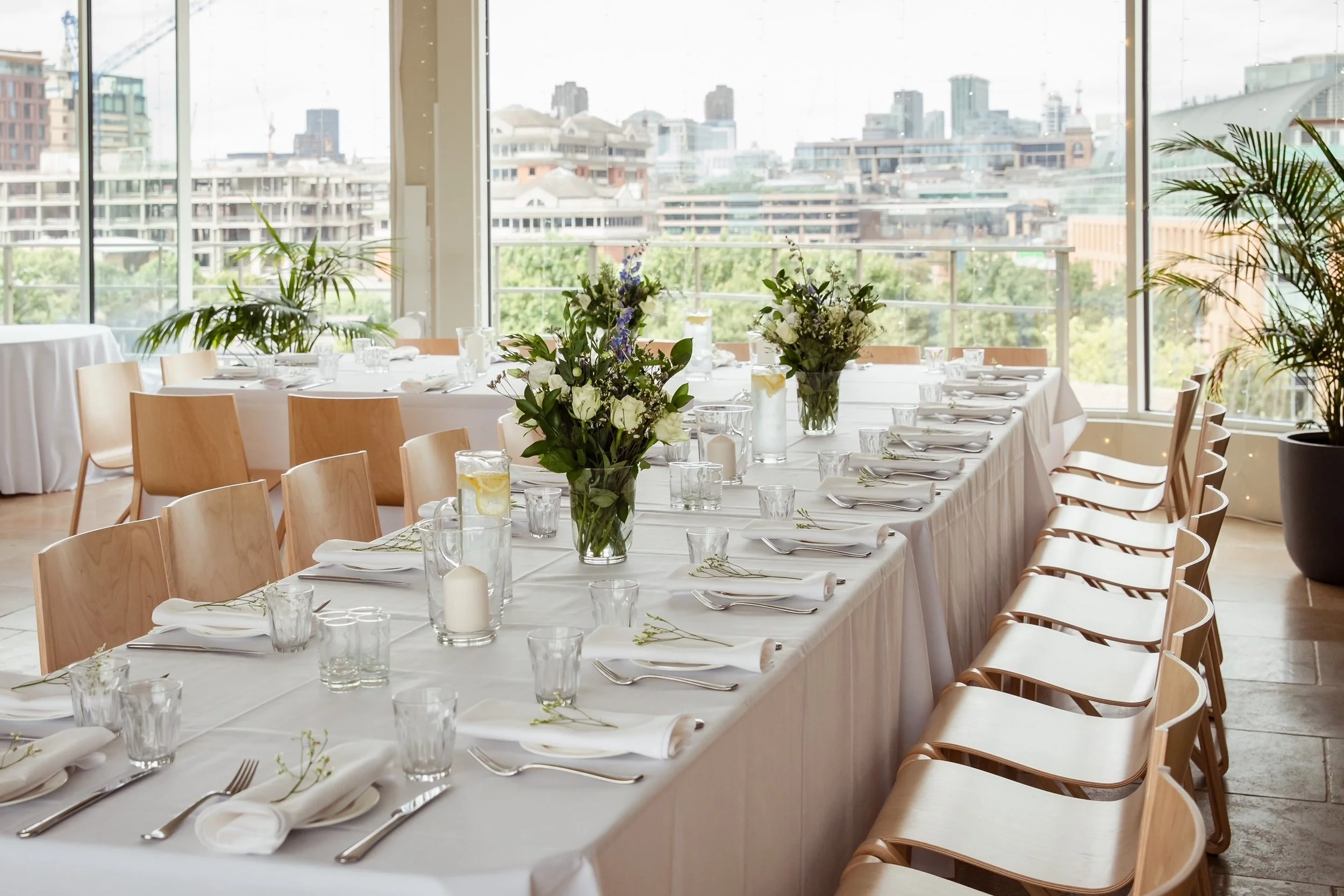 Elegant banquet table set with white tablecloth, floral centerpieces, cutlery, glasses, and napkins in a bright room with large windows and city views.