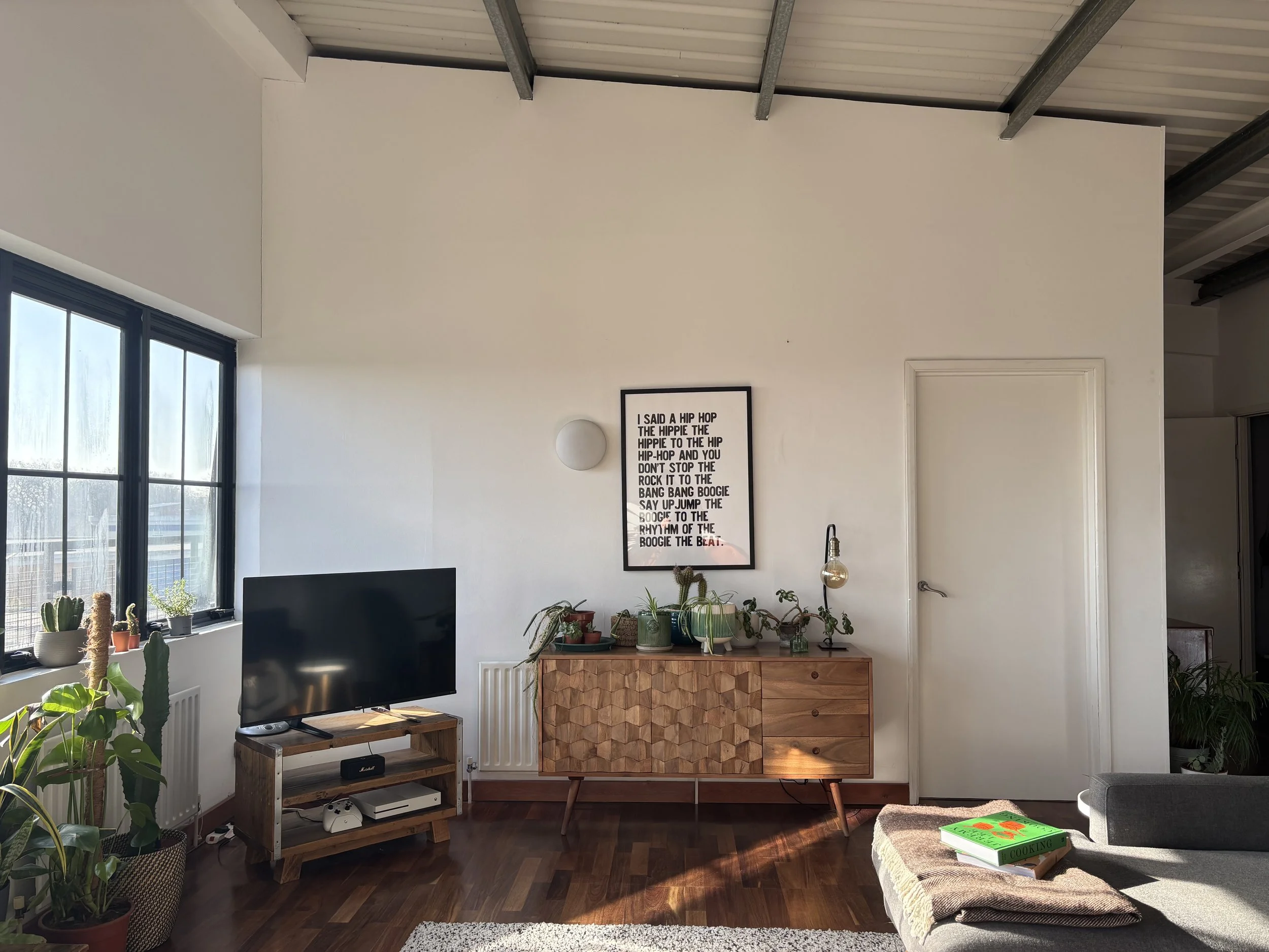 Living room with large window with several potted plants, a flat-screen TV on a wooden stand, a mid-century modern wooden cabinet decorated with plants, a framed print on the wall, and a grey couch with a blanket, book, and pillow.