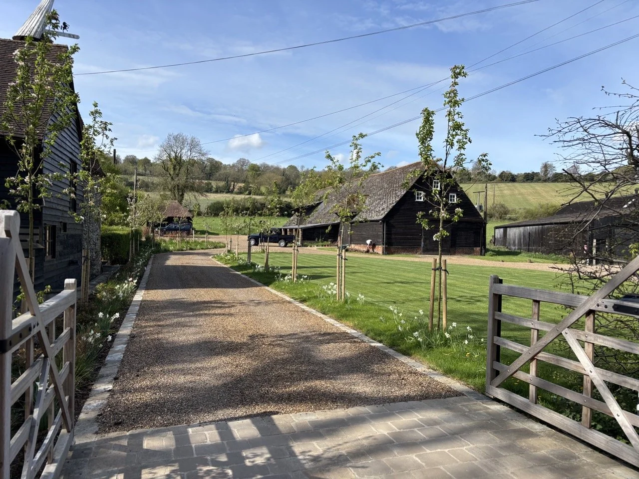 A rural farm scene with a gravel driveway leading to a black barn, surrounded by green grass and young trees, under a blue sky with scattered clouds.