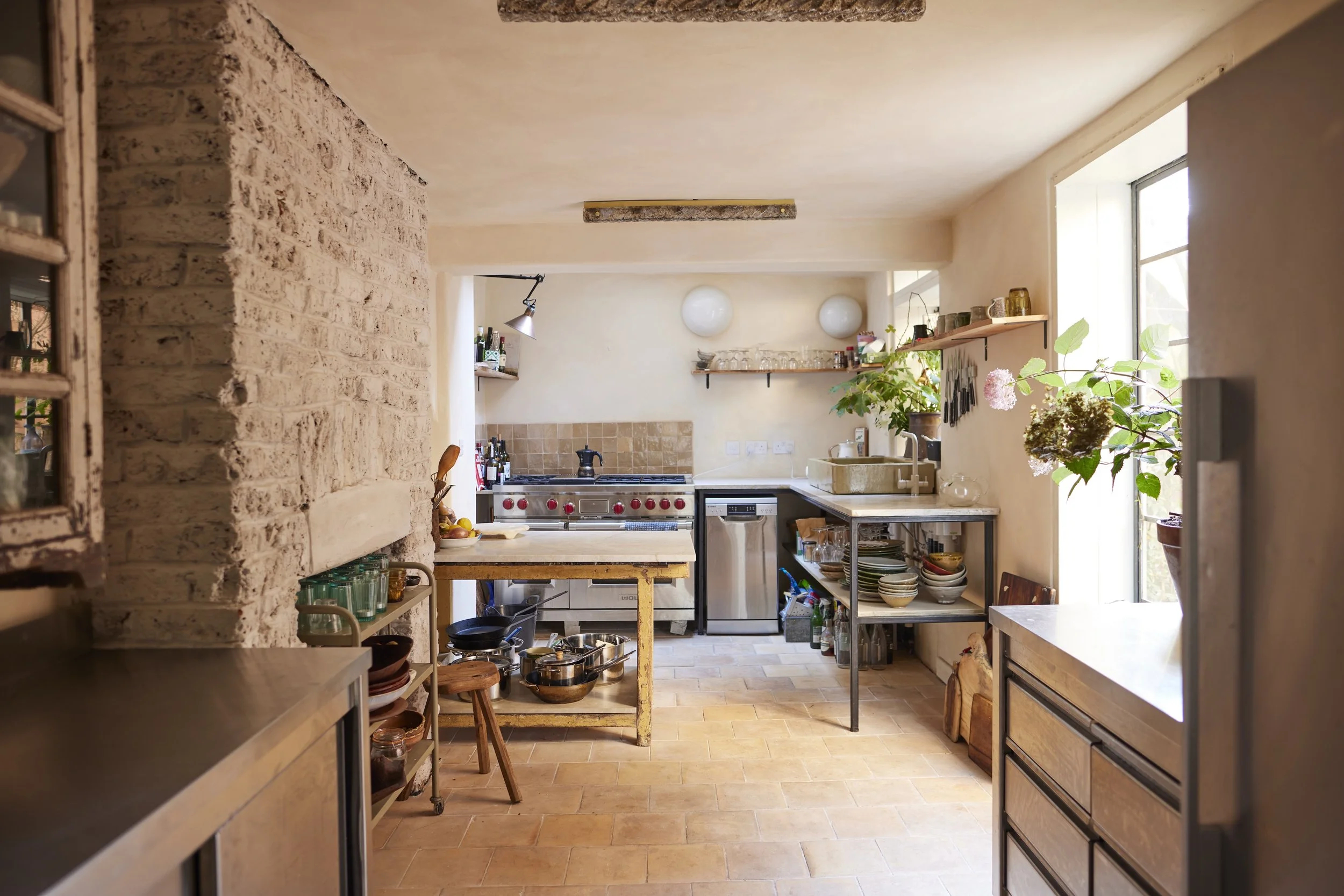 Cozy rustic kitchen with exposed brick wall, wooden shelves, plants, and natural sunlight