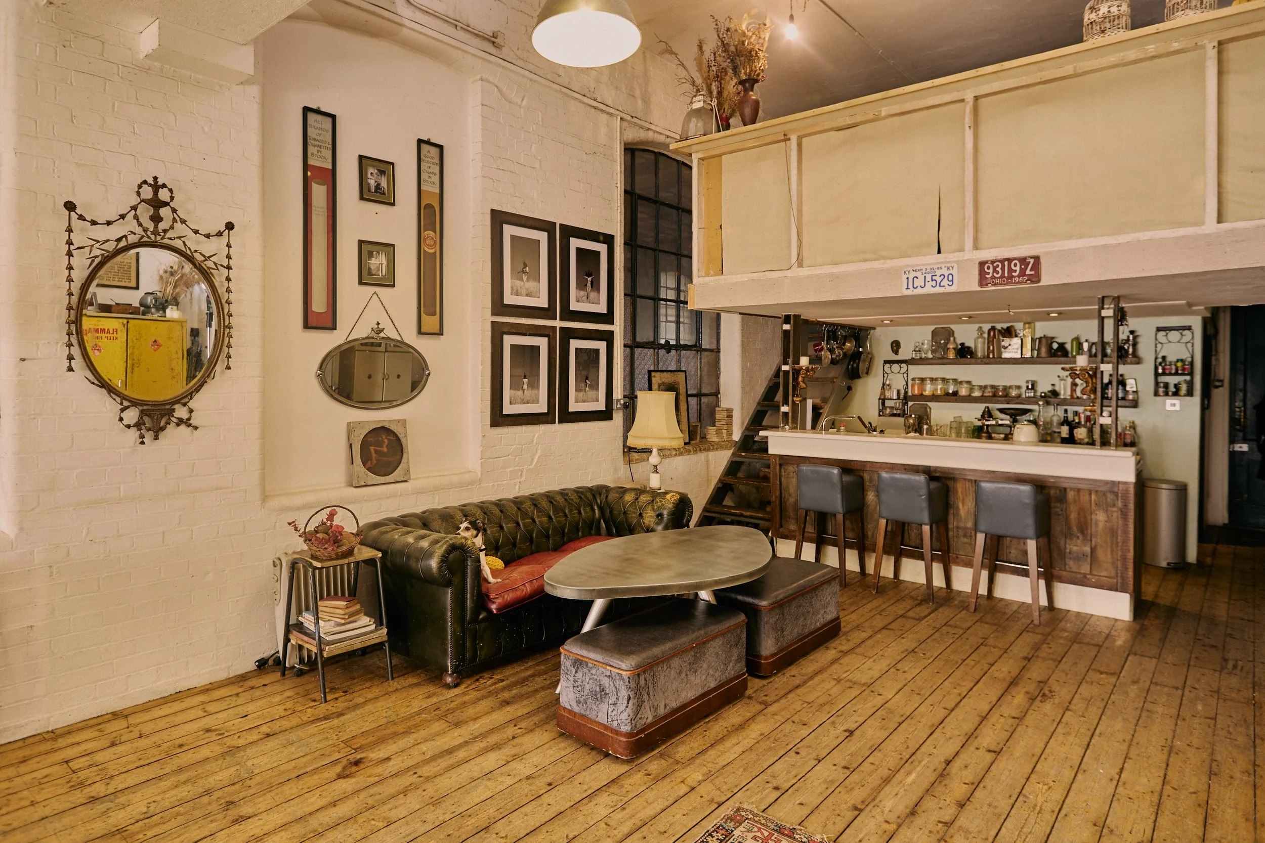 Interior of a cozy cafe or apartment with white brick walls, a leather tufted sofa, a small side table with books and a basket, framed artwork and mirrors on the wall, a wooden bar area with barstools, and a loft with a staircase.