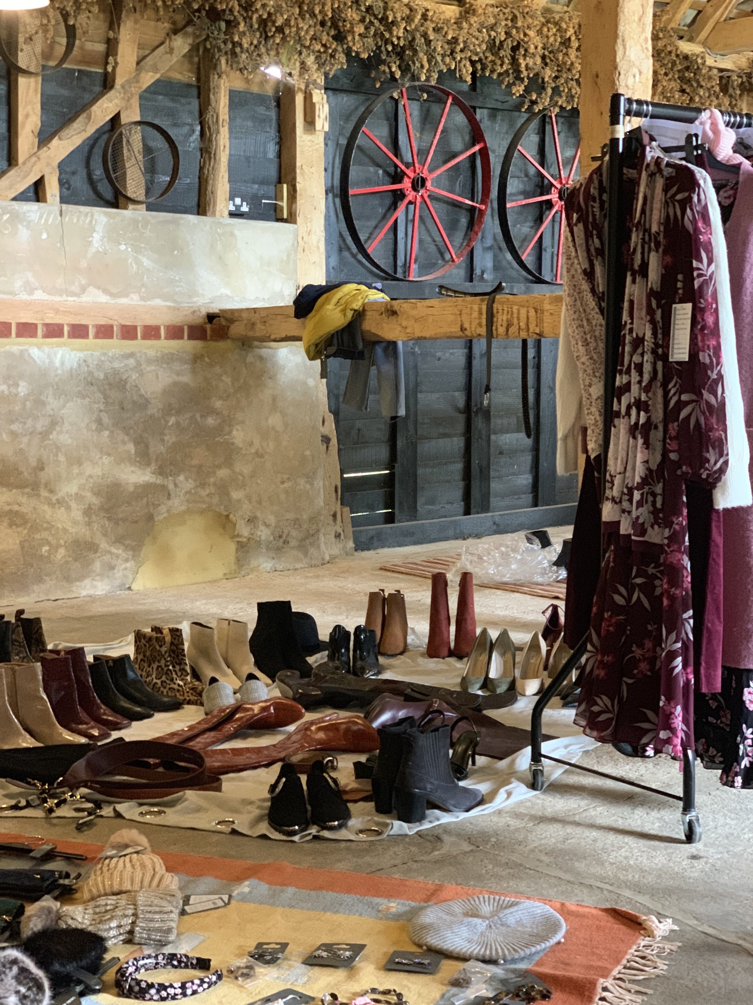 A variety of shoes including boots, heels, and flats are displayed on a white cloth on the floor, with clothing and accessories also visible. The background has wooden and dark panel wall with decorative elements and hanging dried foliage.