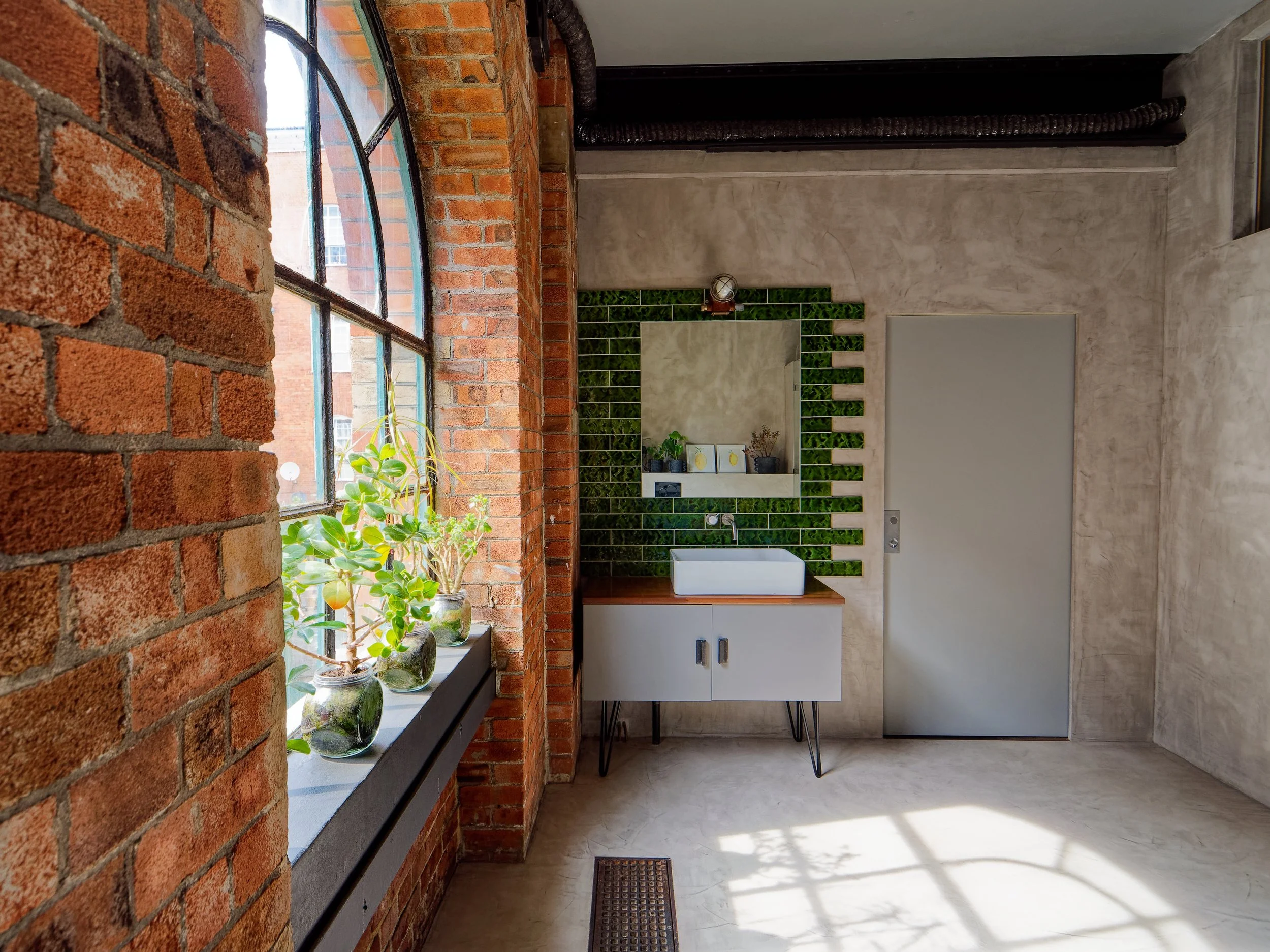 Interior of a modern bathroom with exposed brick walls, a large window with plants on the windowsill, and a vanity with a white sink. There's a mirror framed with green tiles, a closed door, and sunlight casting shadows on the floor.