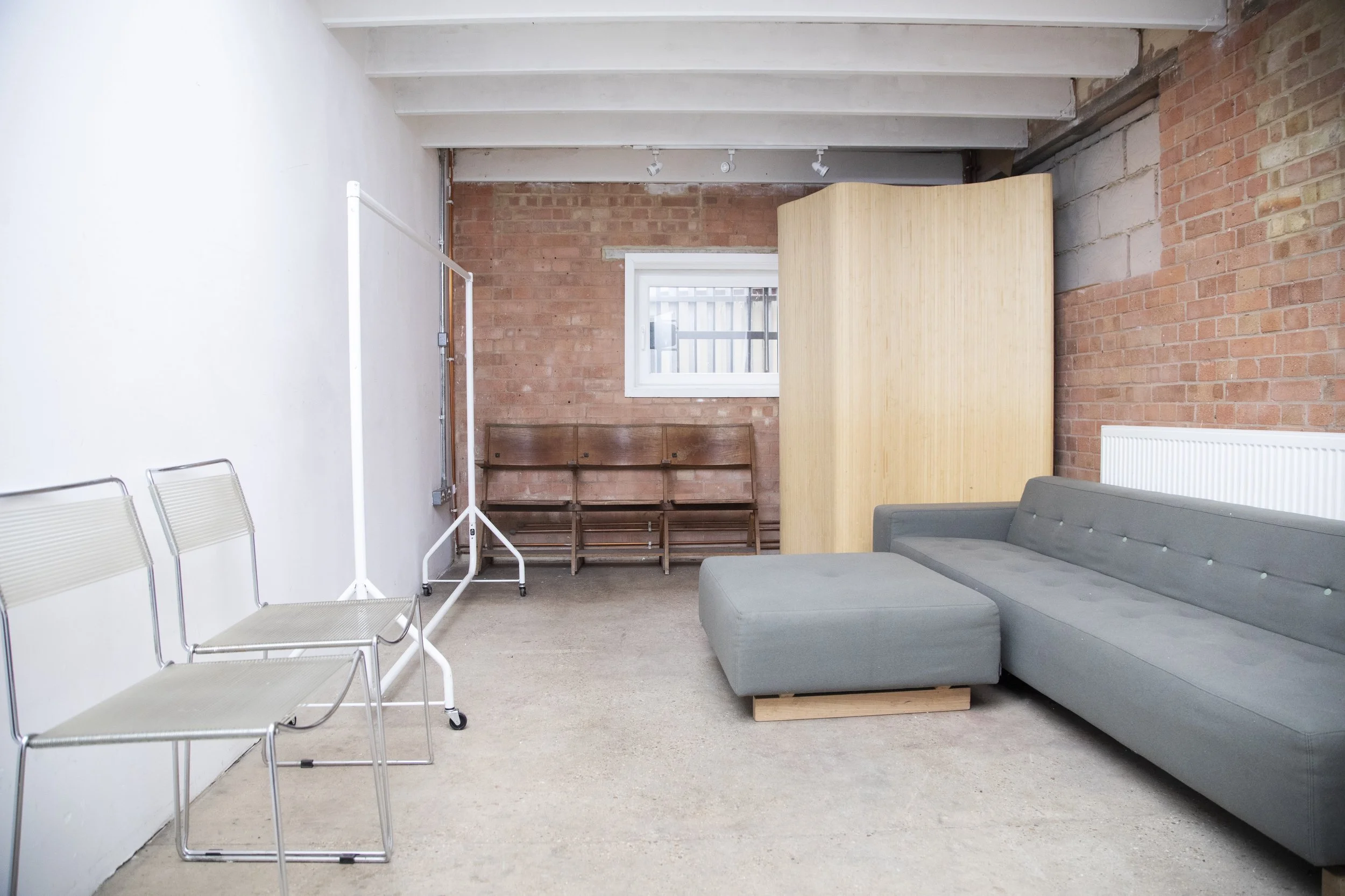 An indoor lounge area with a gray sectional sofa, wooden chairs, a small window on a brick wall, a wooden room divider, and a clothing rack, with a plain white and brick wall background.