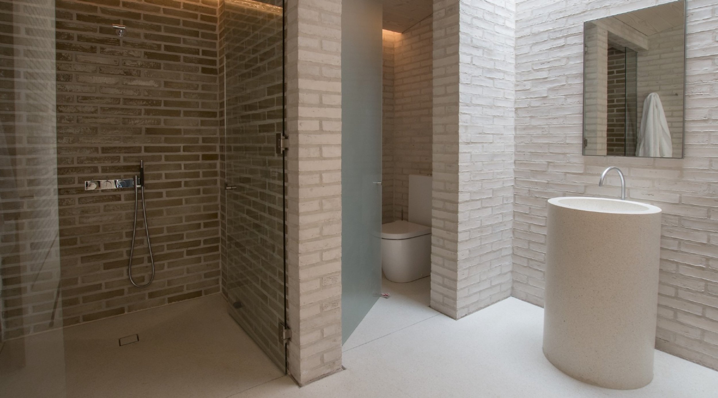 Modern bathroom with a brick shower area, a toilet behind a brick partition, a mirror above a textured pillar sink, and beige brick walls.