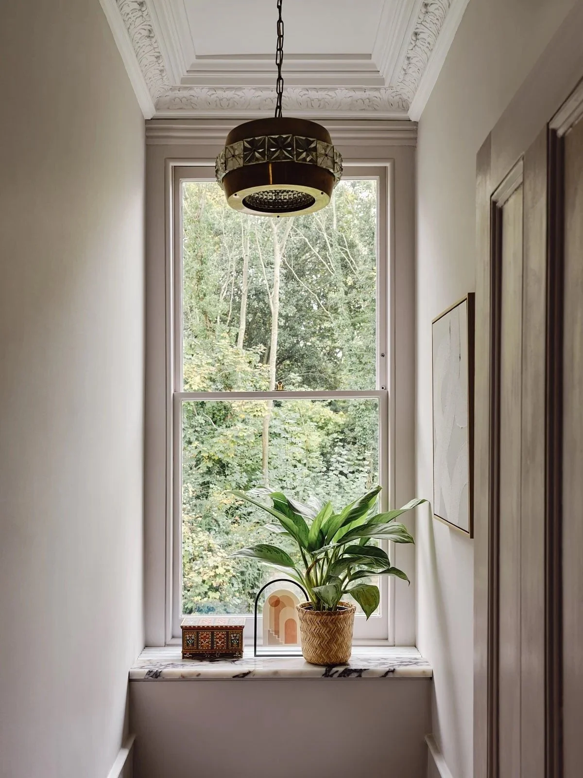 A window ledge with a potted plant, a decorative box, and a small decorative arch sculpture, with a view of trees outside.
