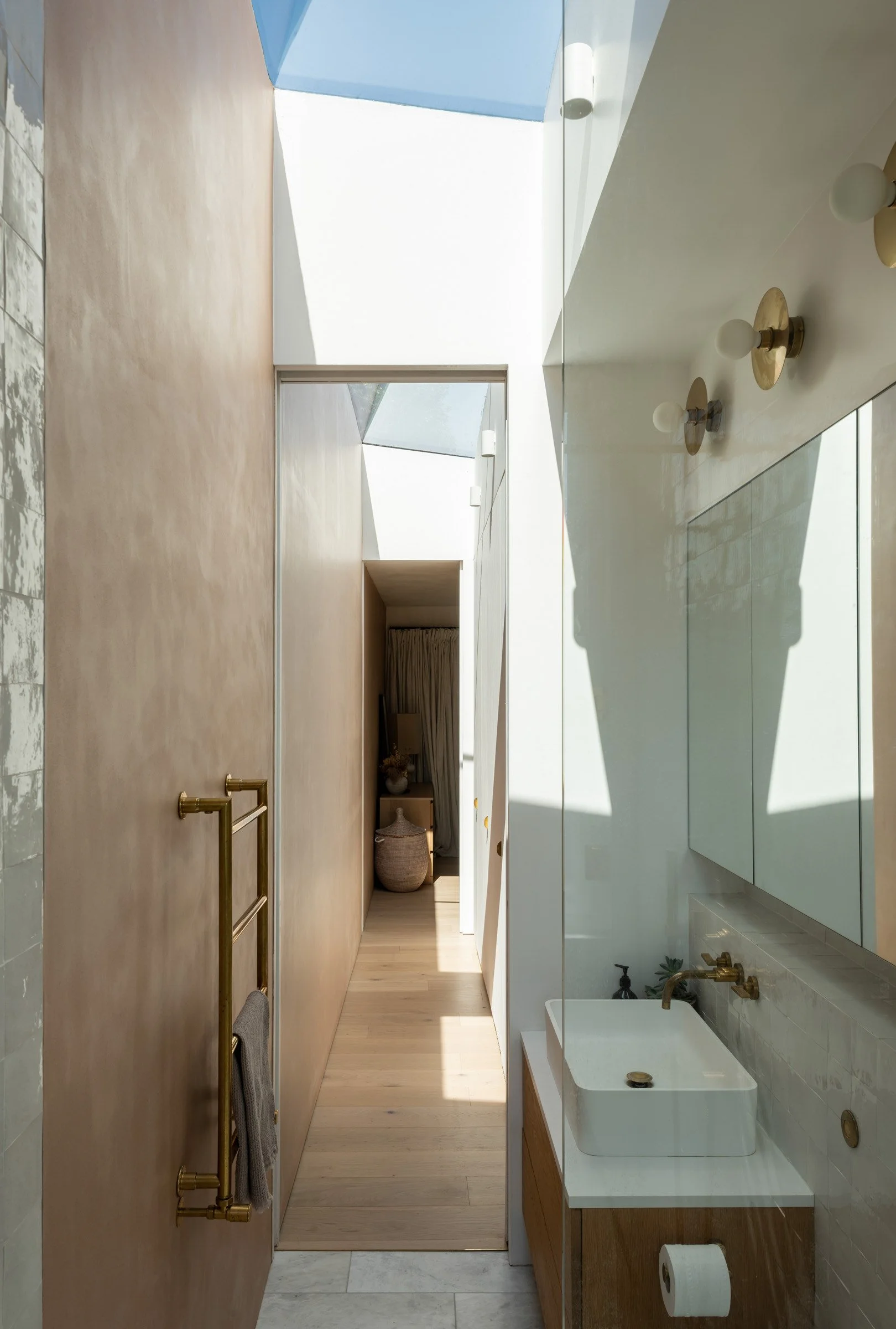 Modern bathroom with a white vessel sink, gold fixtures, a large mirror, and a skylight ceiling with sunlight and shadows.