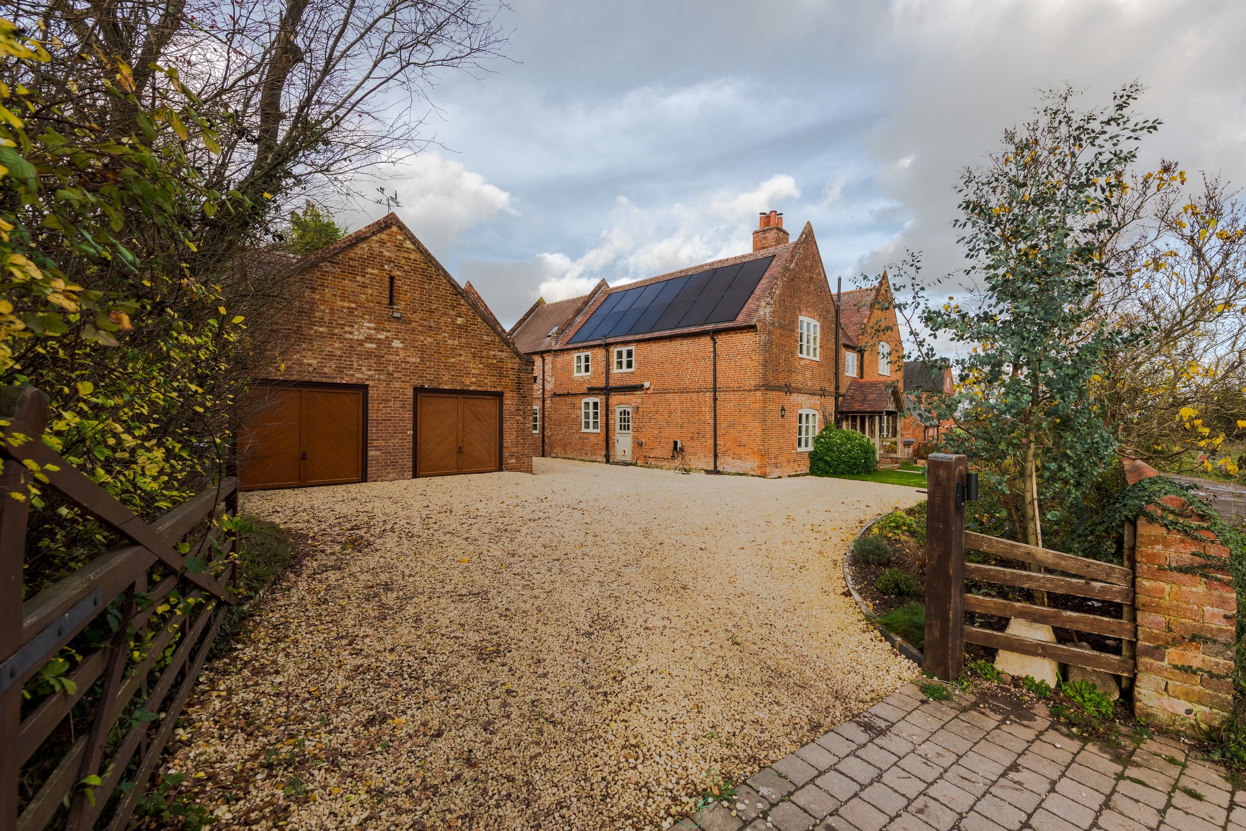 A large brick house with solar panels on the roof, situated in a gravel driveway with a wooden gate and surrounding trees and bushes.