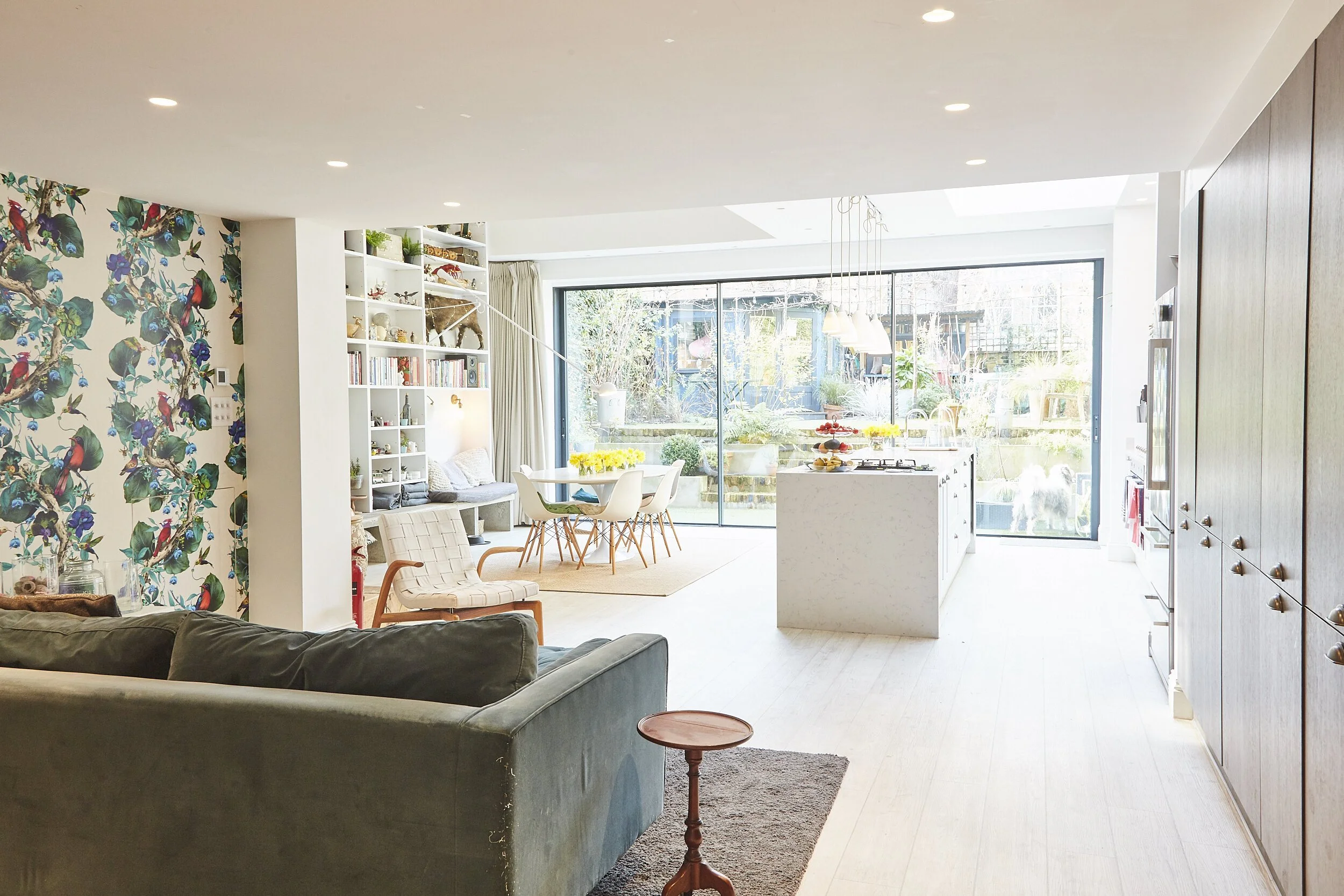 Bright open-plan living area with a view of a backyard, featuring a sofa, a small wooden side table, a dining table with chairs, and a kitchen island with food. Large glass sliding doors lead to the outdoor garden, which has various plants and outdoo