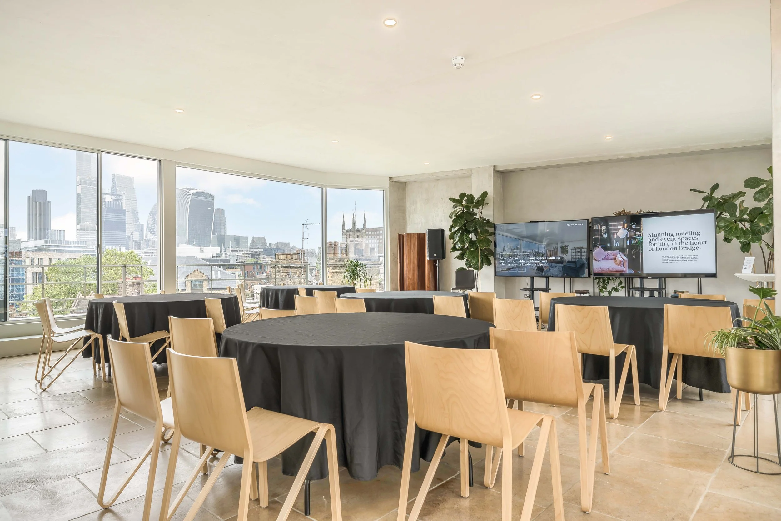 A bright, modern conference room with large windows showcasing a city skyline, round tables covered with black tablecloths, and wooden chairs. There are two large screens displaying a presentation and some tall plants for decoration.