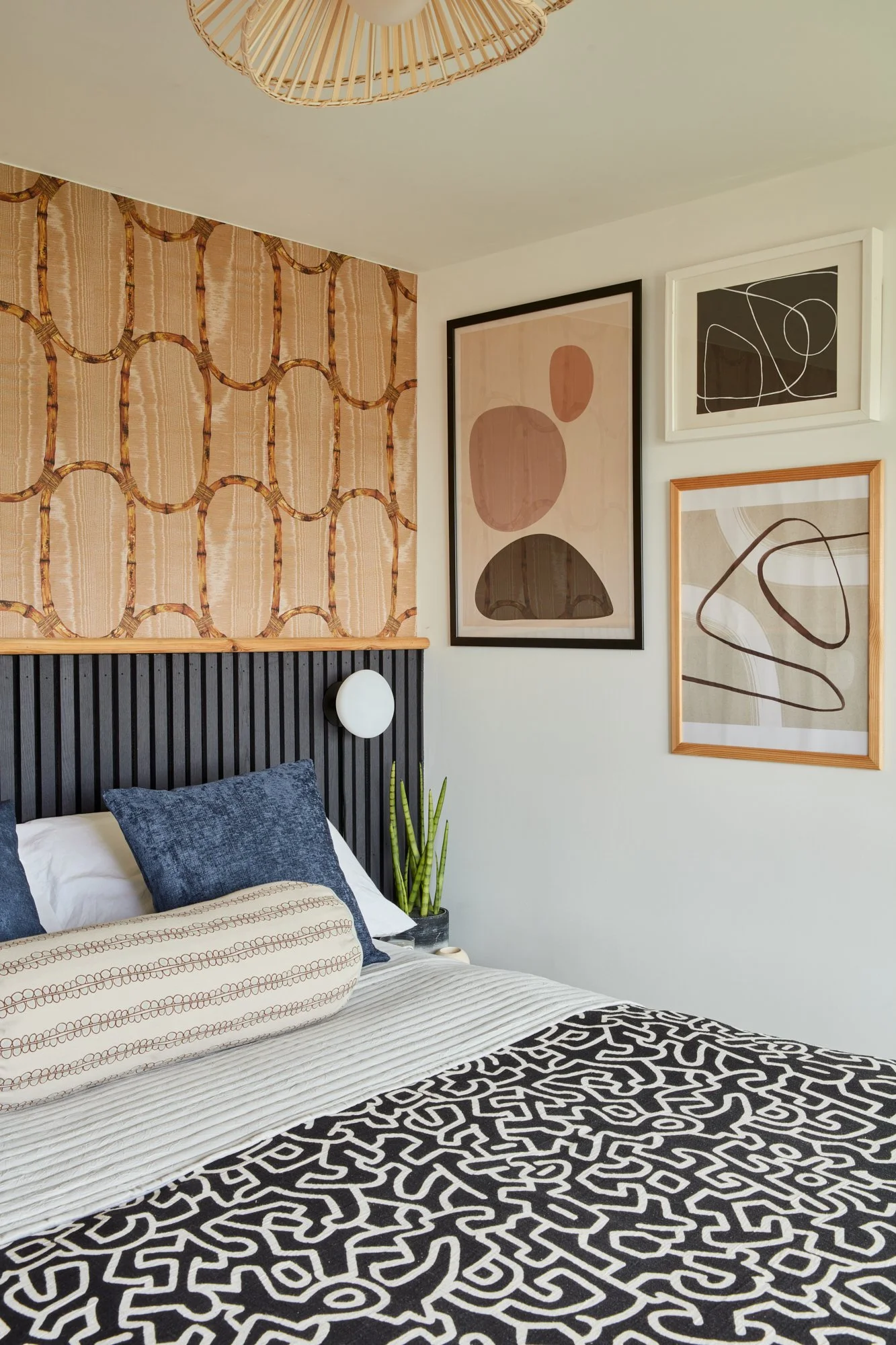 A bedroom featuring a black wooden headboard, a white bed with dark blue pillows, a patterned bench at the foot of the bed, and a wall adorned with three abstract art prints. There is a small plant on the bedside table next to the headboard and a wov