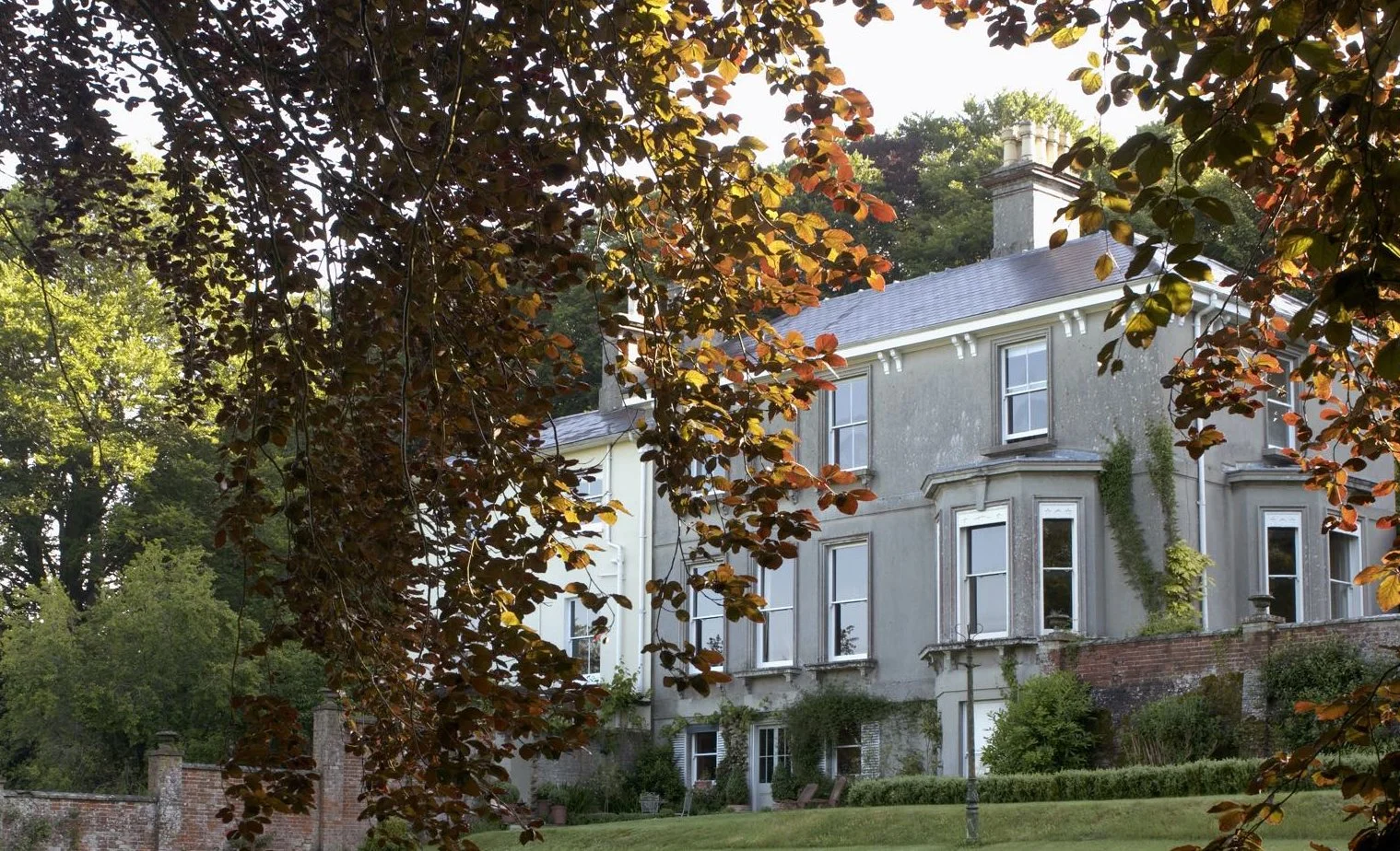 A large, old mansion with a grey exterior, multiple windows, and a chimney, surrounded by trees with green and reddish leaves.