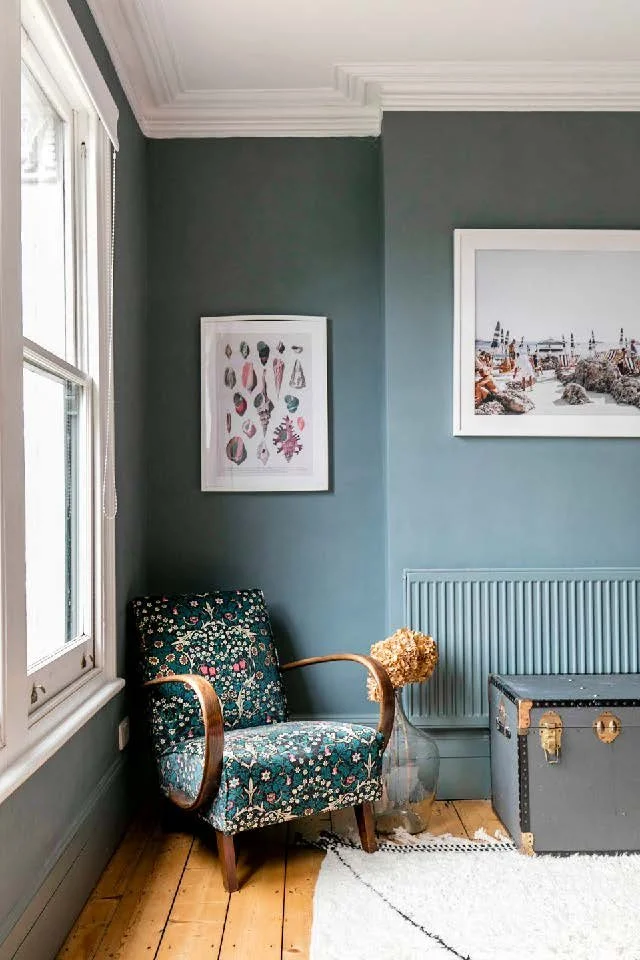 A cozy corner of a room with a patterned armchair, a glass vase with dried flowers, a chest, and framed artwork on blue walls.