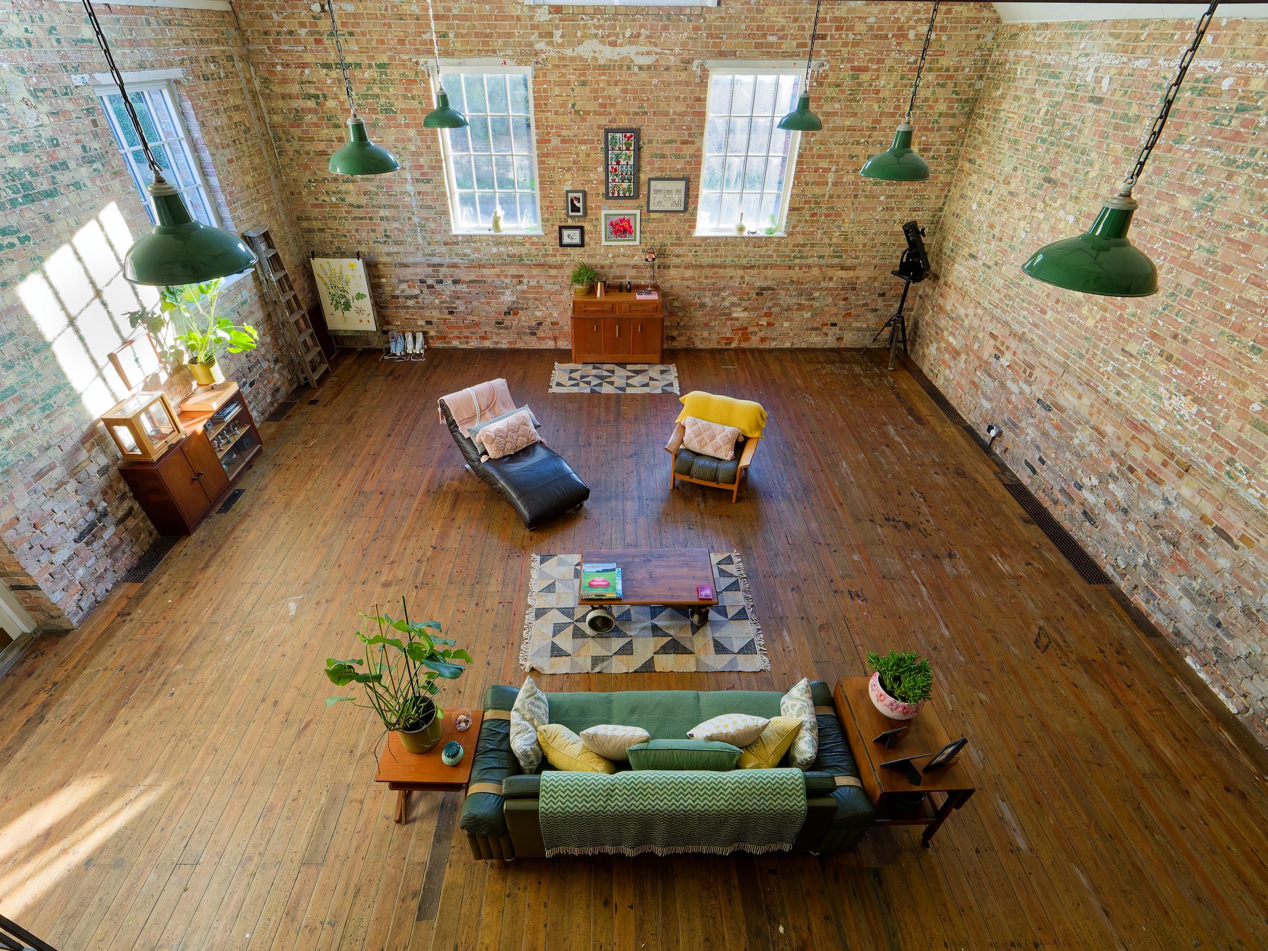 A spacious loft with brick walls, wooden floor, and large windows. It has a green leather couch with yellow and white pillows, and a coffee table on a geometric rug. There's a black lounge chair with a pillow, a wooden side table with plants and deco