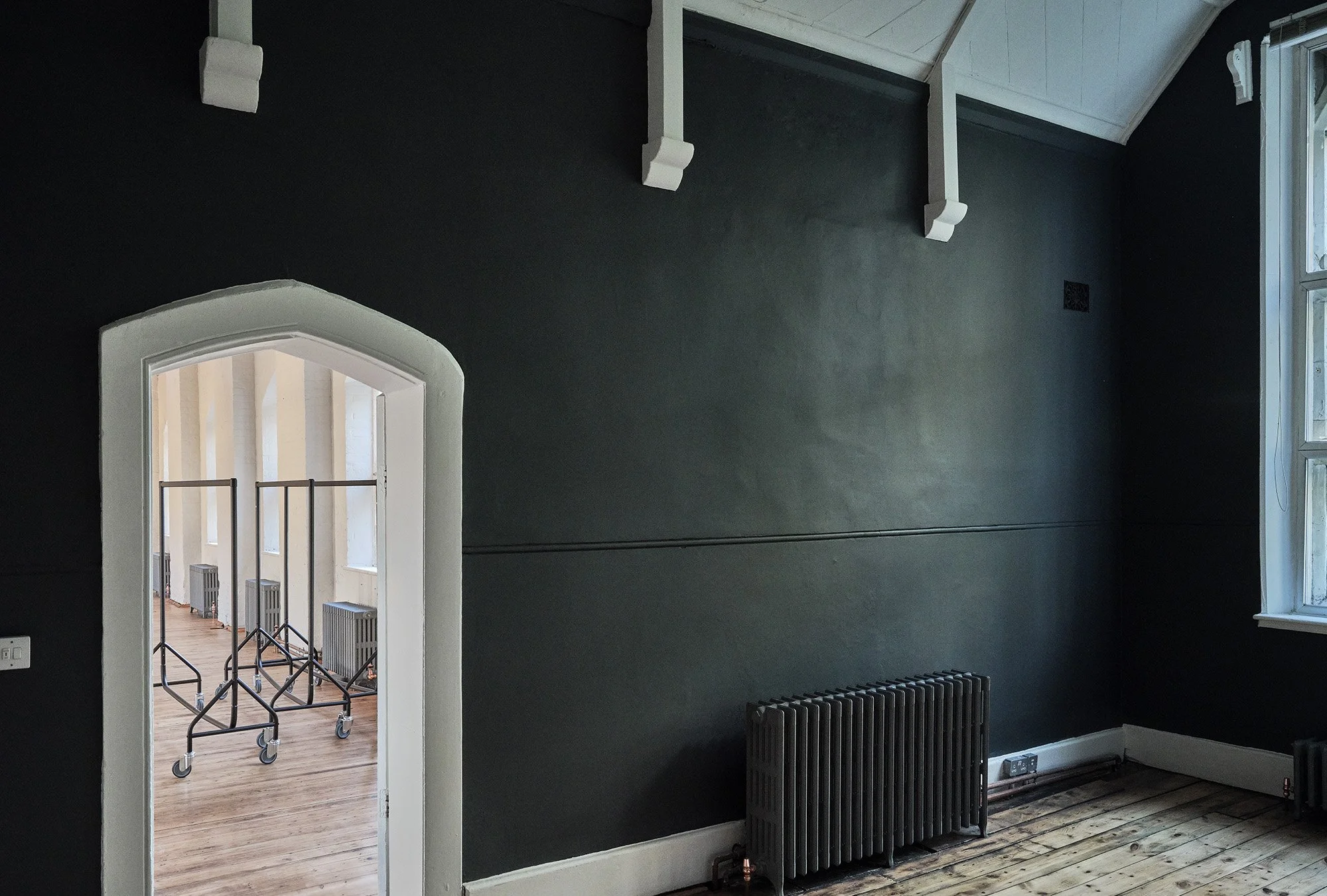 Empty room with dark painted wall, high ceiling with white beams, window letting in natural light, and a vintage radiator on the wall.