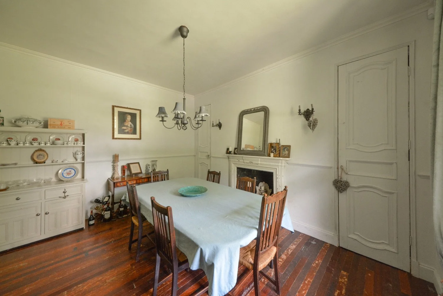 A dining room with a wooden table covered with a white tablecloth, six wooden chairs around it, a white china cabinet filled with dishes, a framed portrait hanging on the wall, a mirror above a fireplace, and vintage decorative items and wall sconces