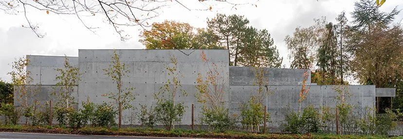 A modern, minimalist concrete wall with small trees and shrubs planted along its base, set against a cloudy sky and autumn foliage.
