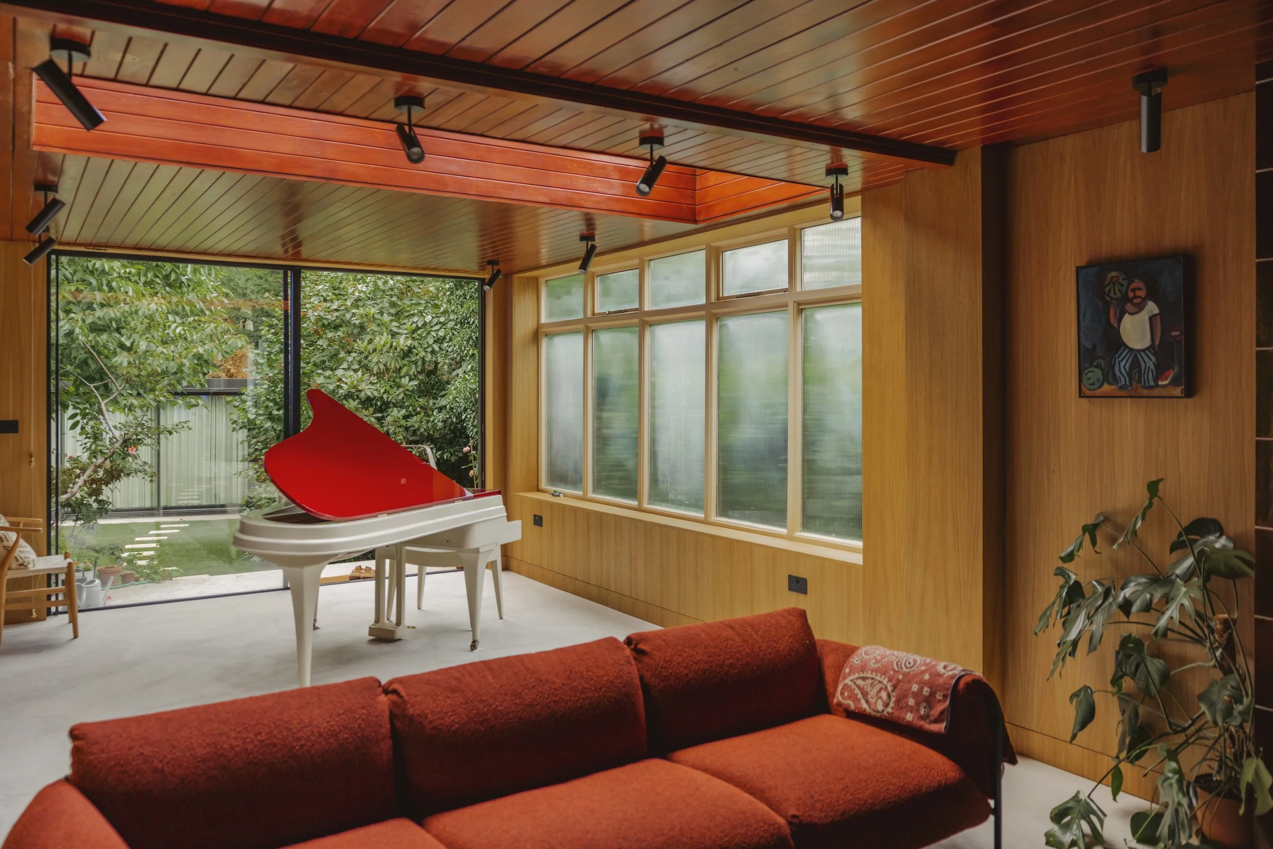 Living room with wooden walls and ceiling, large windows, a white grand piano with a red lid opened, and a red sofa in the foreground.