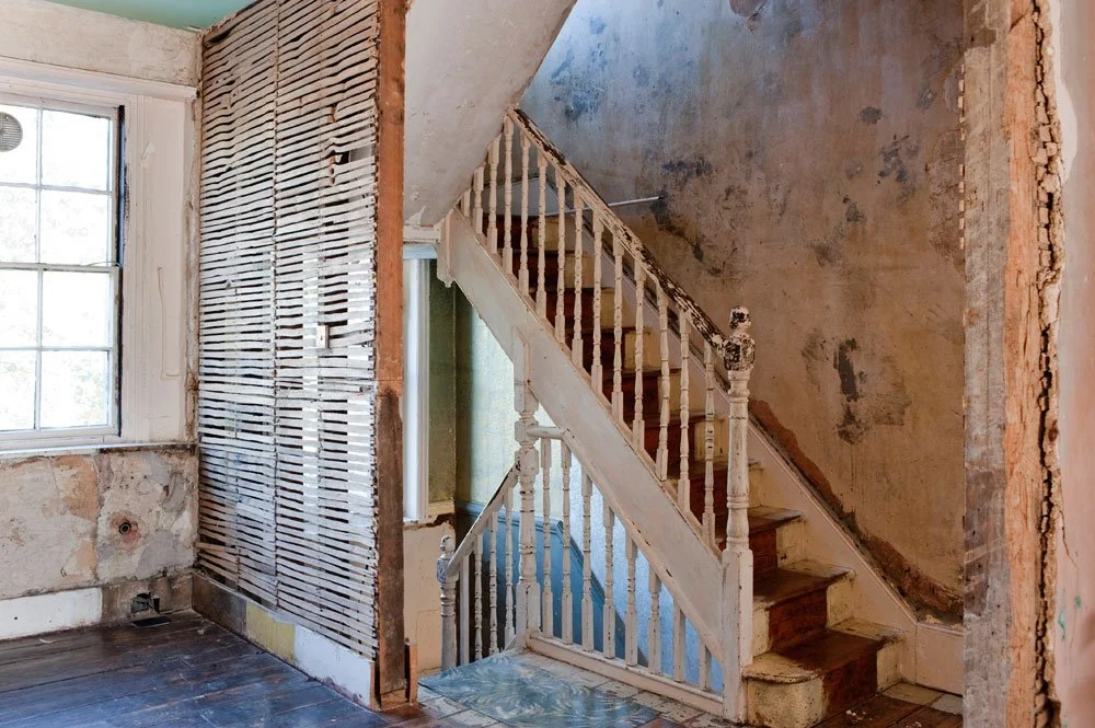 Interior of an old house with peeling paint, exposed walls, and a worn staircase with chipped white banisters.