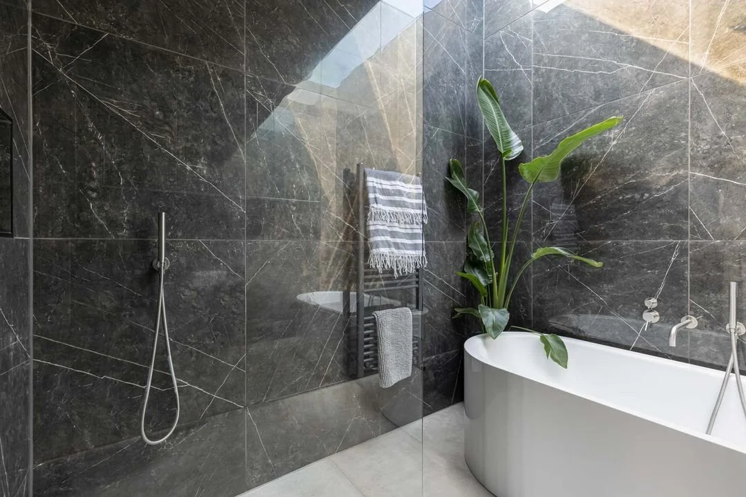Modern bathroom with black marble walls, a white bathtub, and a large green plant near the tub. Towel rack with striped and gray towels, and a handheld showerhead on the wall.