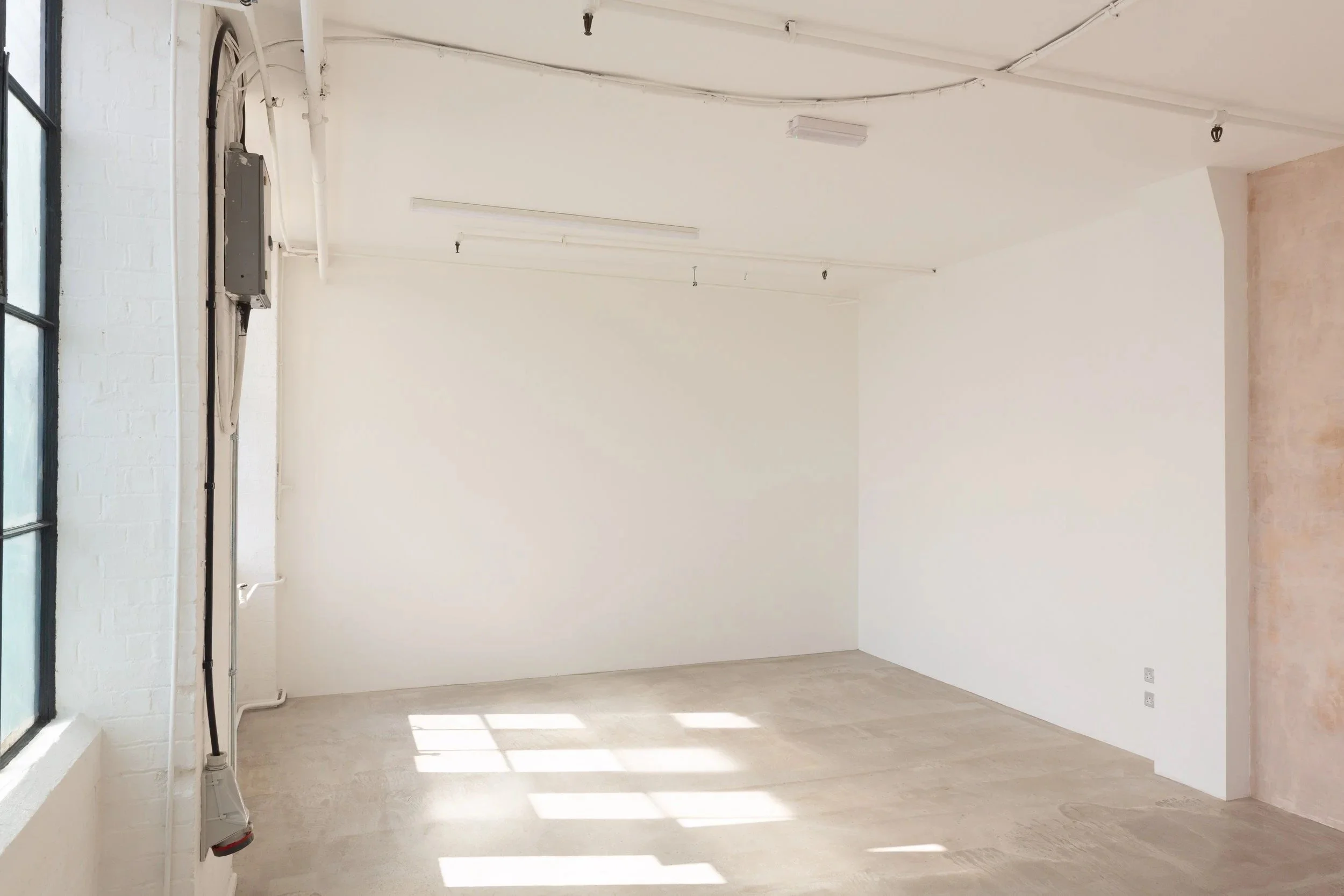 Empty room with white walls, large window on the left, sunlight casting shadows on the floor, unfinished wall on the right, exposed electrical fixtures on the ceiling.