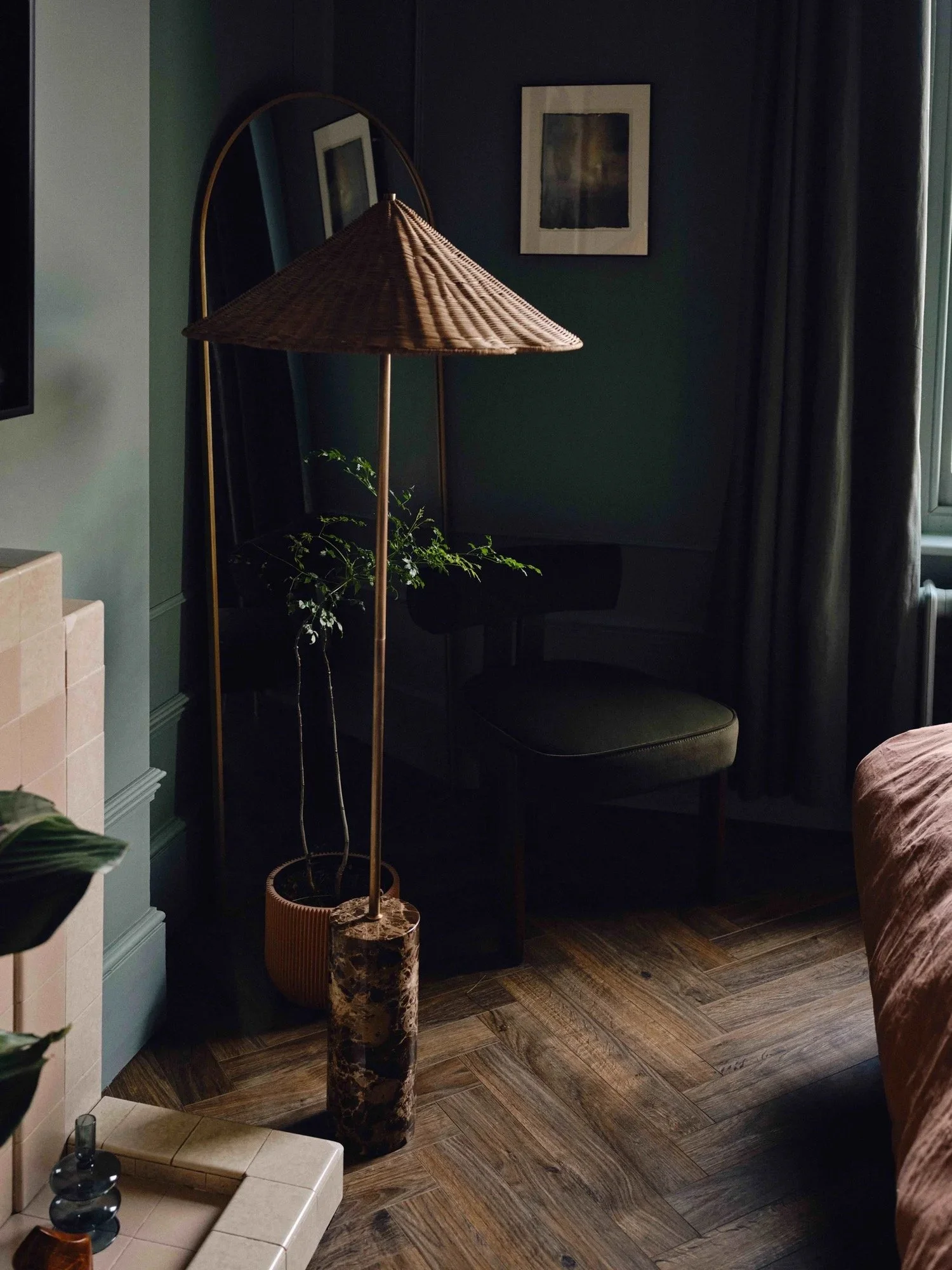Interior of a room with a green wall, a window with curtains, a mirror, framed artwork, a dark chair, a floor lamp with a woven shade, a potted plant, and a tiled surface.