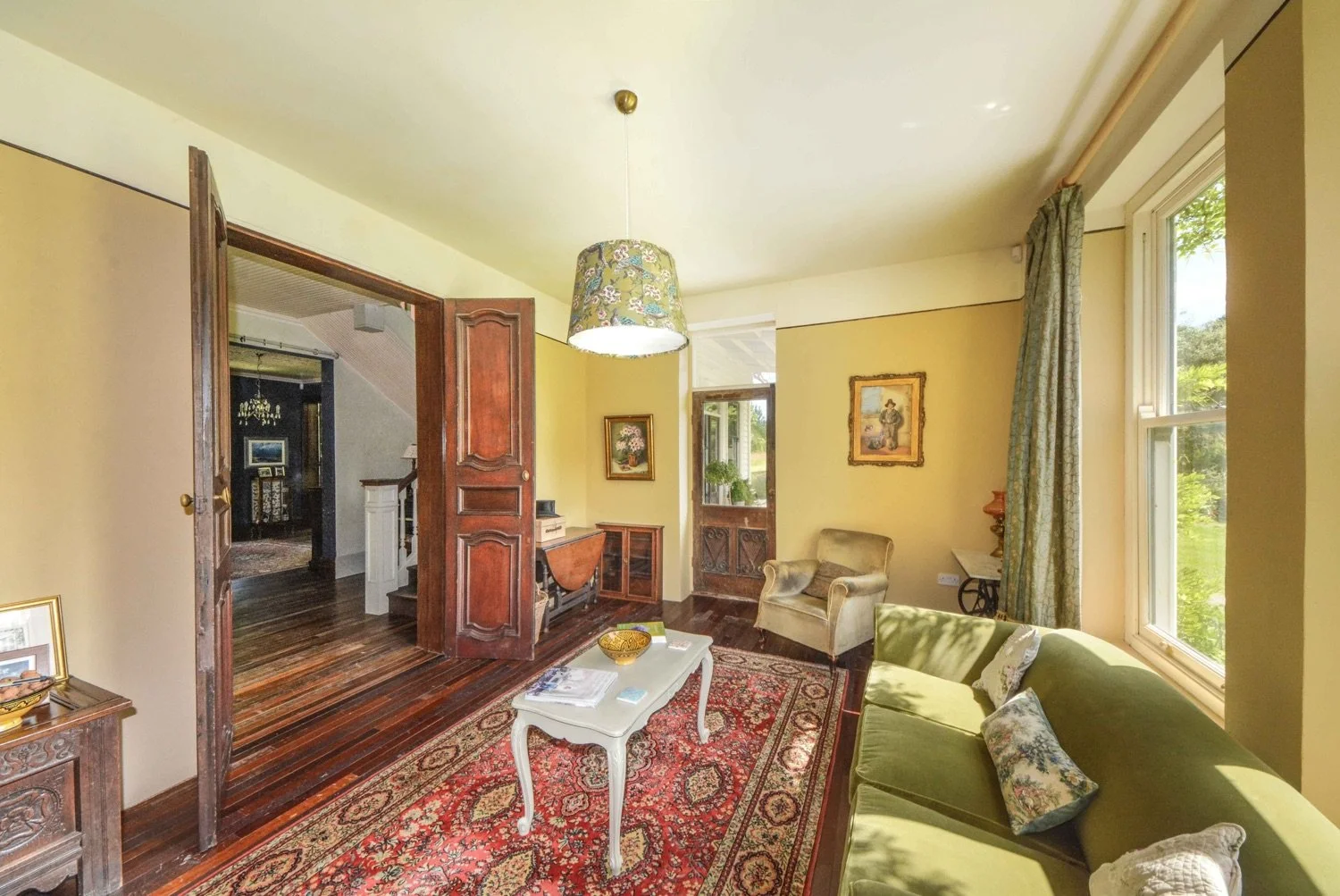 Bright living room with yellow walls, large windows with curtains, green sofa, beige armchair, wooden doorways, a white coffee table, and red patterned rug. Decor includes paintings and a floral lampshade.