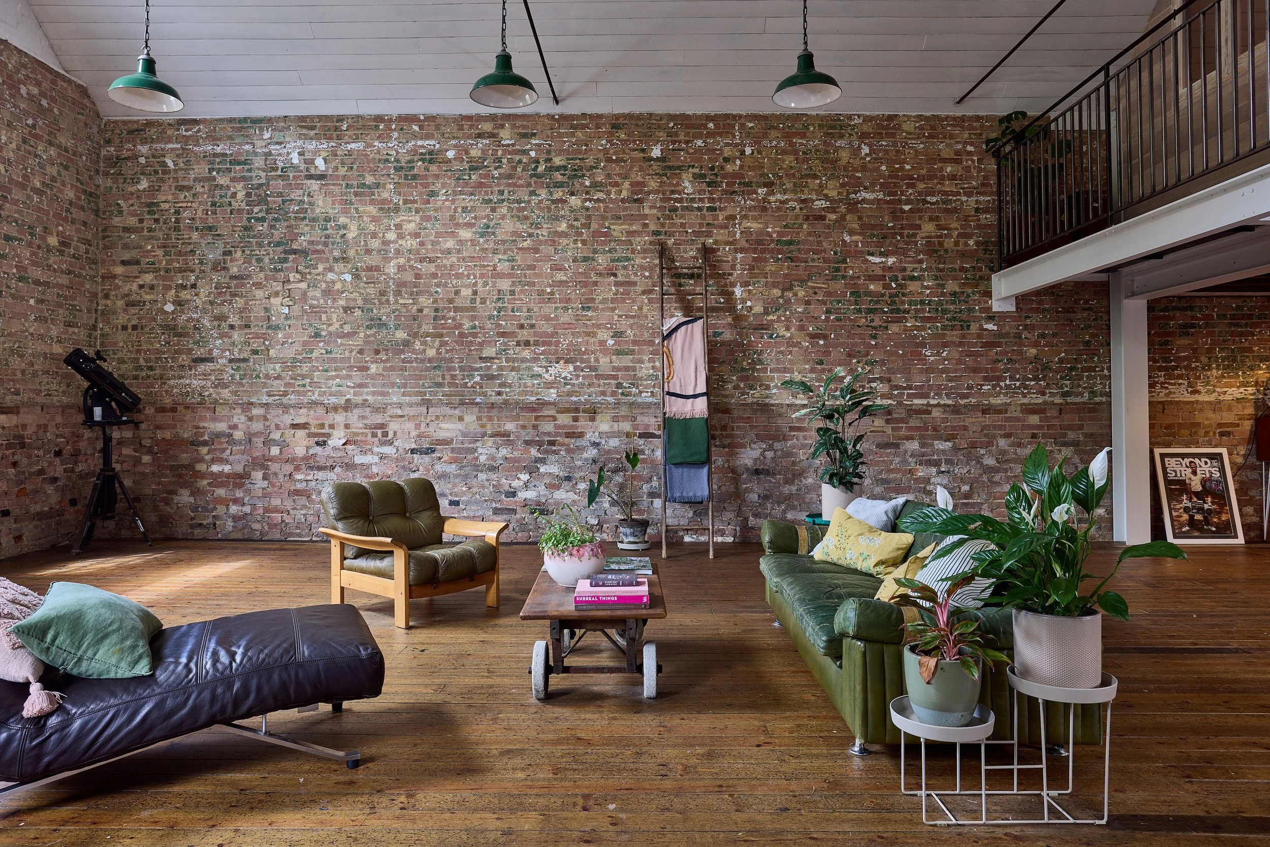 Living room with exposed brick wall, wooden floor, and ceiling. Furniture includes a black leather lounge chair with pillows, a green leather sofa with pillows, and an armchair. There are several potted plants and a wooden table with books. A telesco