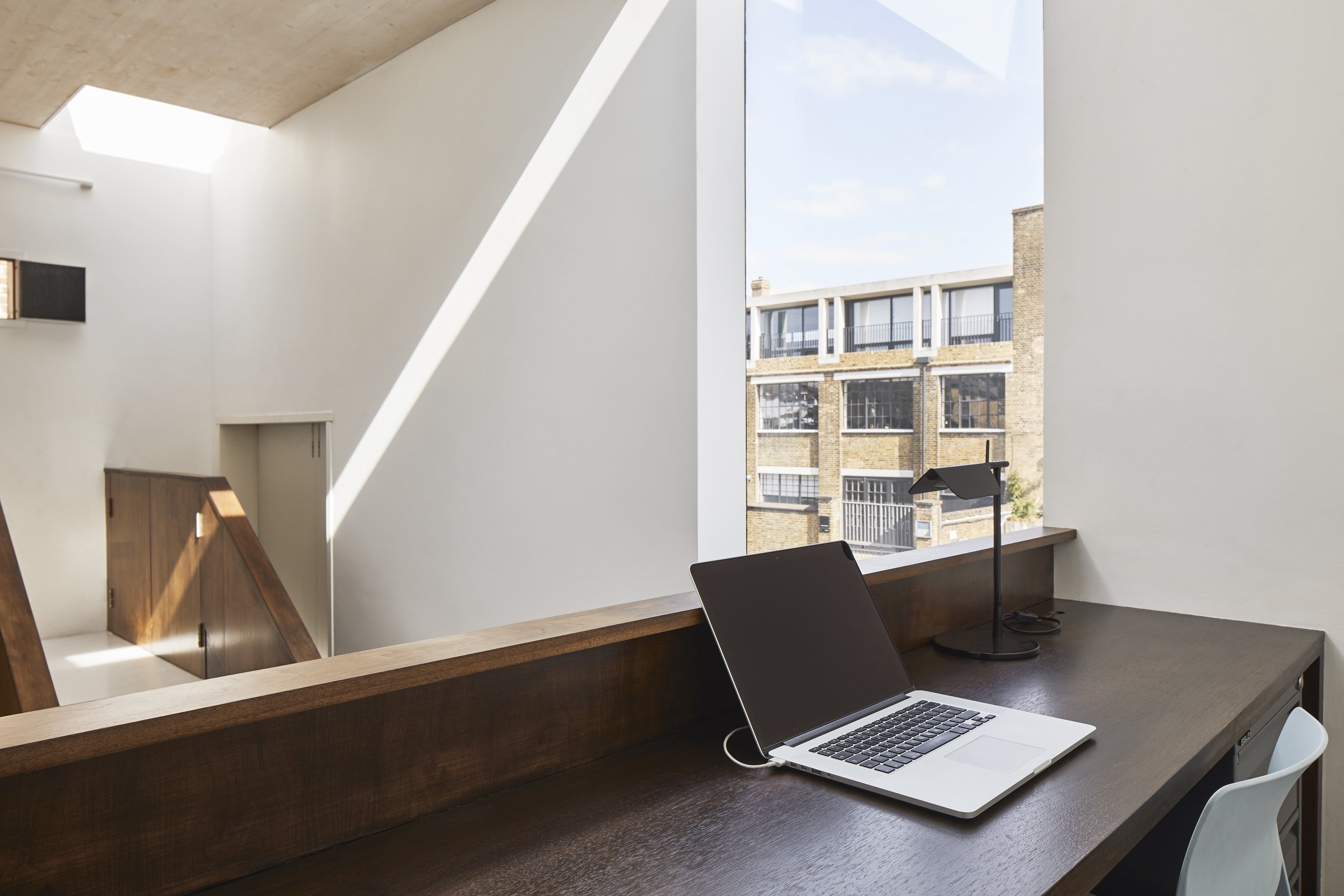 Interior shot of a modern workspace with a wooden desk, an open laptop, a desk lamp, and a white chair, with a view of a brick building through a large window.