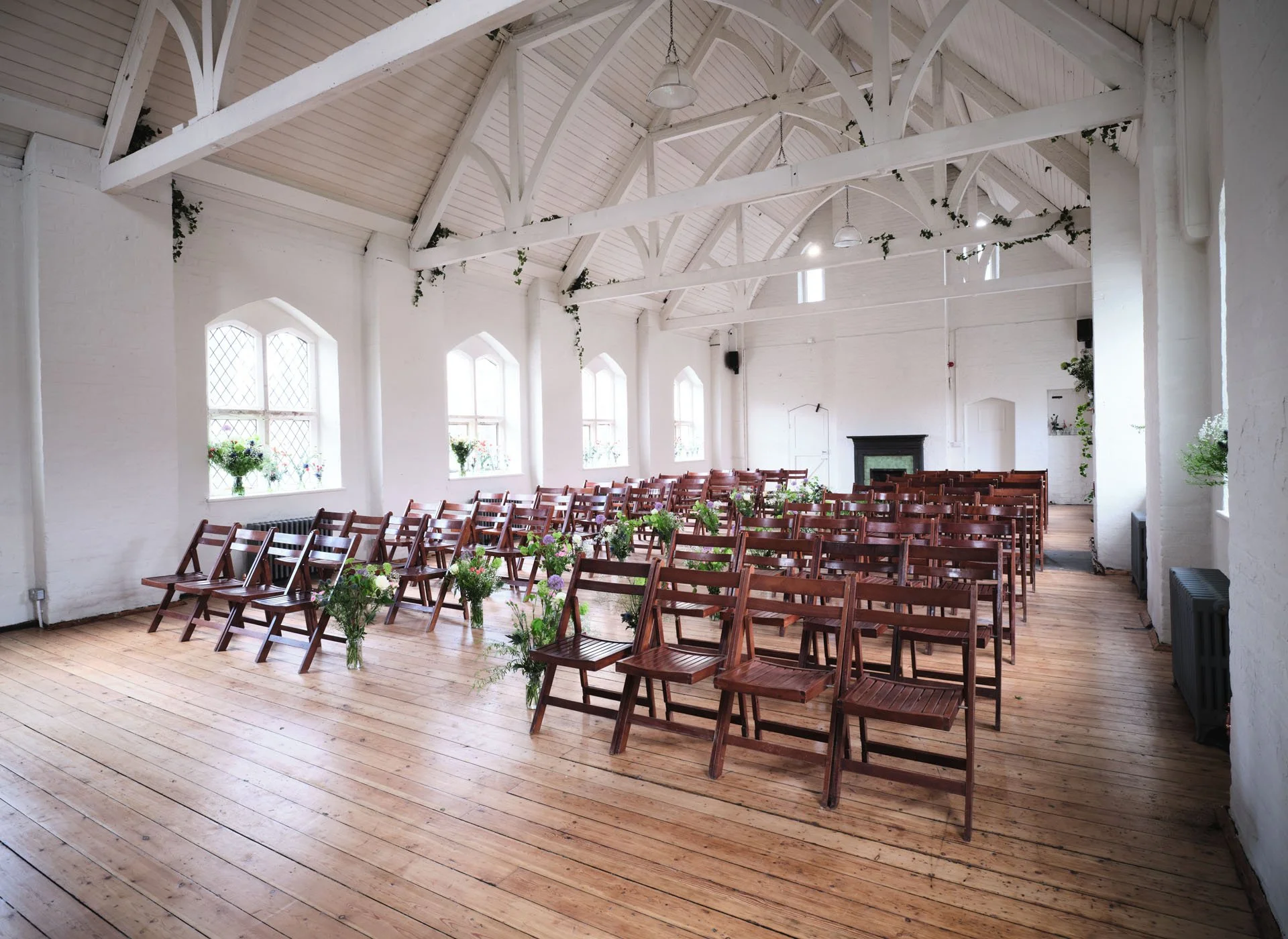 Empty event space with white walls, wooden flooring, rows of wooden chairs, and floral arrangements, decorated for a wedding or ceremony.