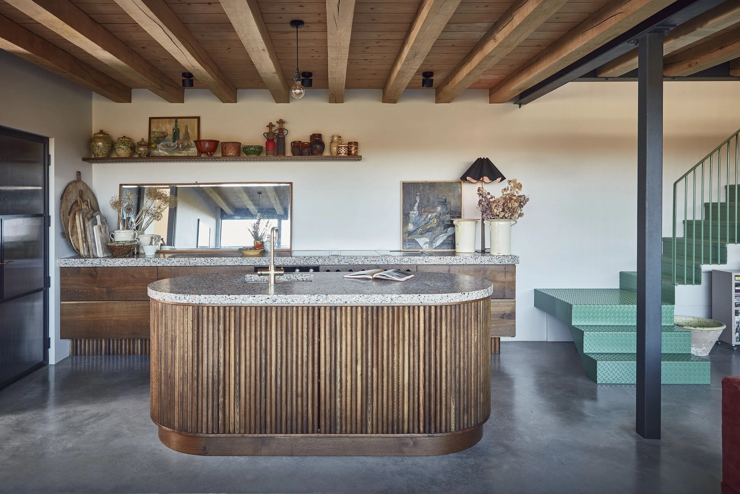 A modern kitchen with a wooden ceiling and an island with a speckled countertop. Decor items like vases, artwork, and a black lampshade are on the wall. Green stairs are visible on the right.