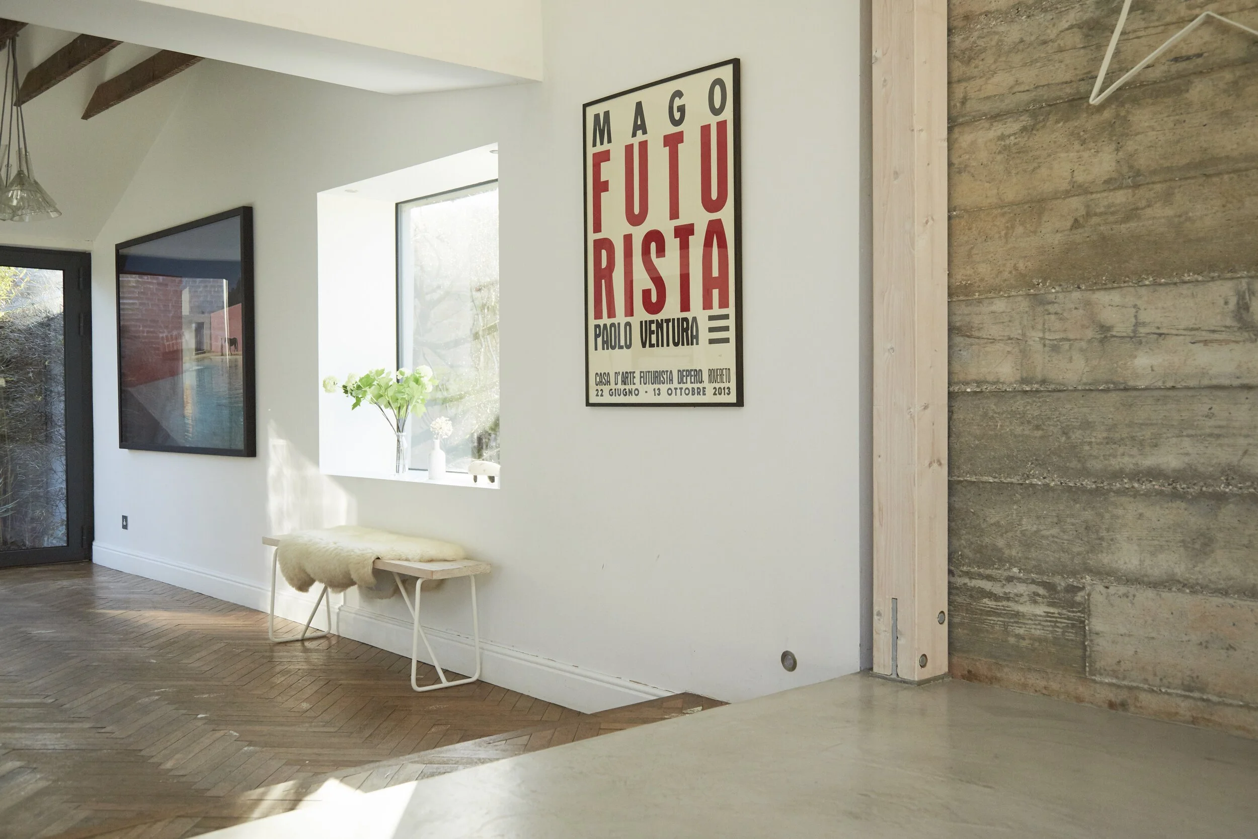 Interior of a modern room with a white wall, large windows, a framed poster with text in Italian, a small bench with sheepskin, a potted plant, and a wooden accent wall.