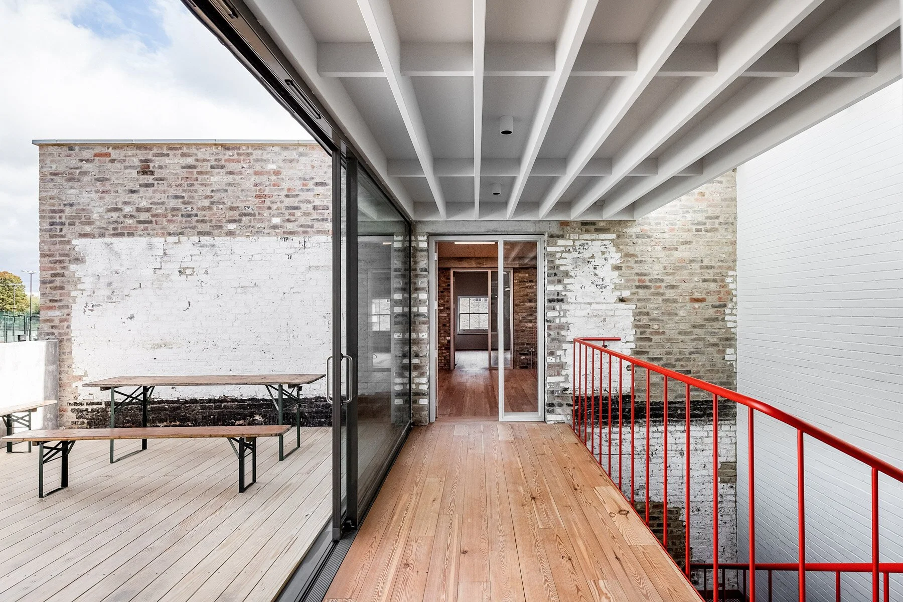 View of a modern balcony with wood flooring, red railing, and a glass sliding door leading to an indoor space. The background shows a brick wall with some white paint, and a wooden table with benches on the outside terrace.