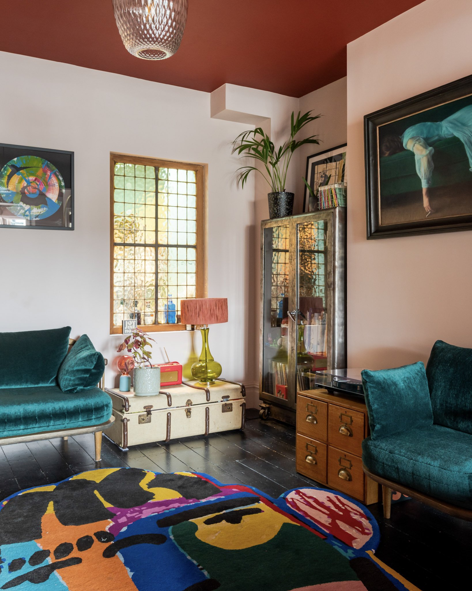 Living room with teal velvet sofas, colorful abstract rug, vintage trunk with a pink table lamp and plants, glass display cabinet, and framed artwork on white and pink walls with a window.