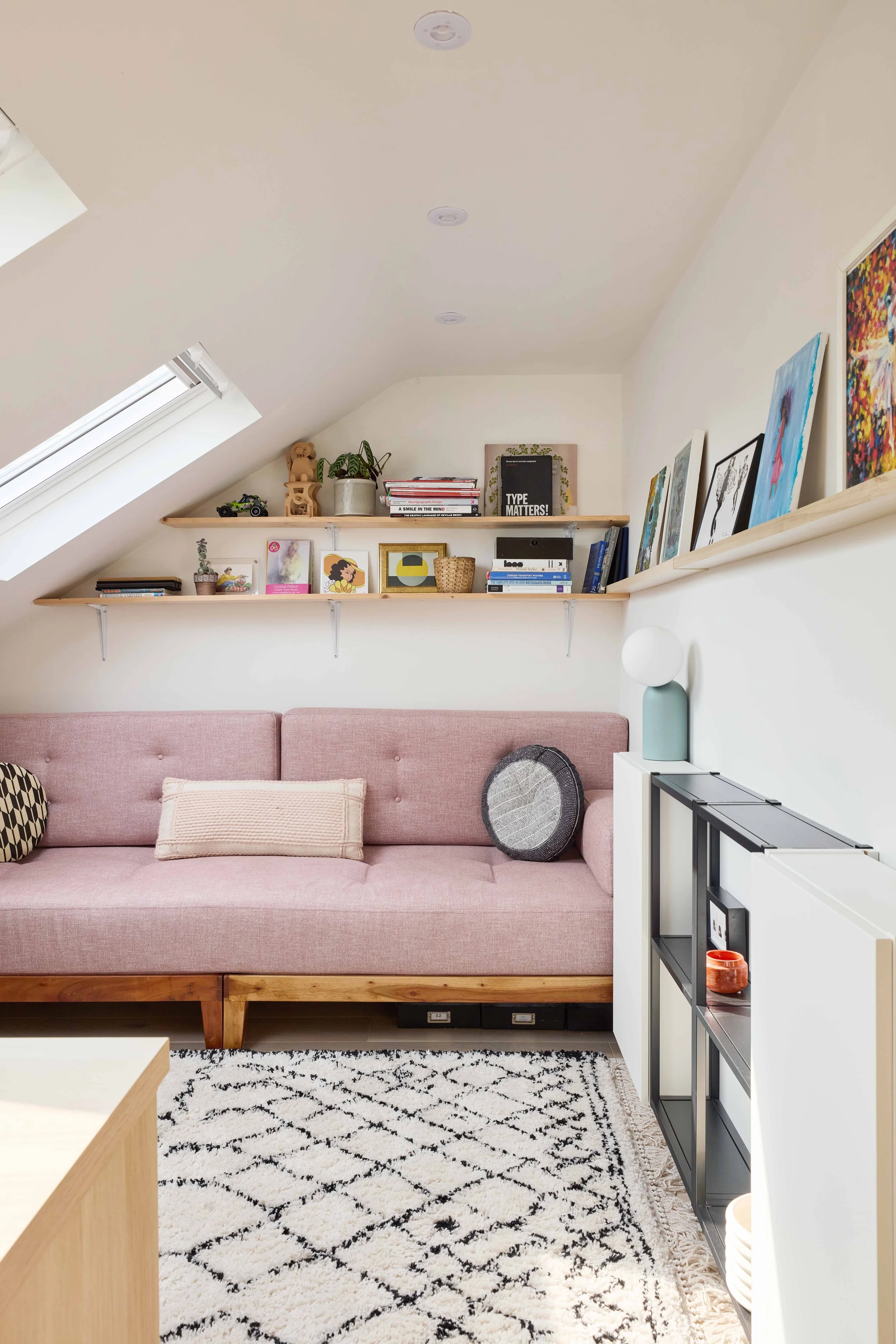 A cozy living room with a pink couch, a white and black patterned rug, and two wooden shelves filled with books and decorative items on a white wall. There is a skylight window on the left side and a black metal shelf on the right side.