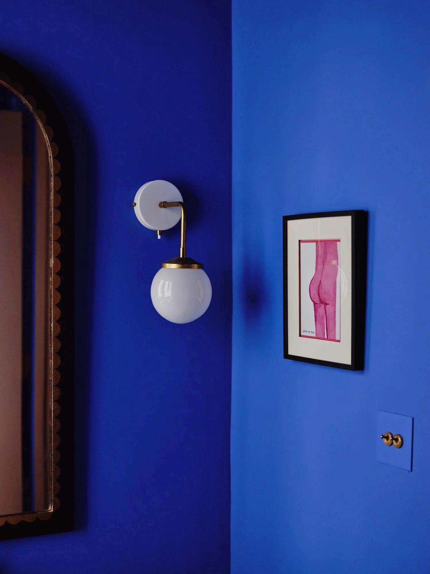 A blue wall with a framed pink art piece, a mounted wall sconce with a white globe shade and brass accents, and a partially visible mirror with a dark brown scalloped frame.