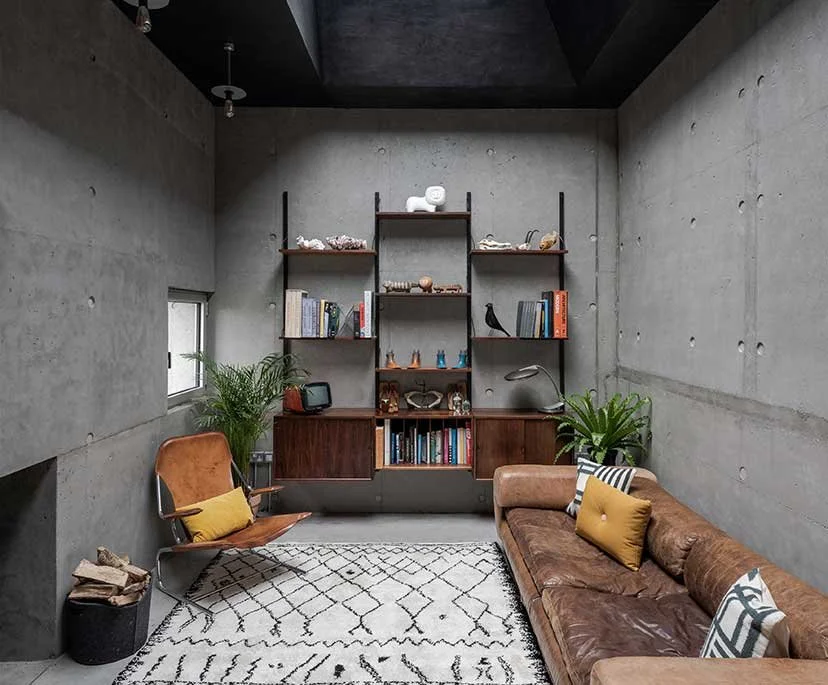 A modern living room with concrete walls, a brown leather sofa with decorative pillows, a rust-colored armchair with a yellow pillow, a black and white patterned rug, two green potted plants, and a tall wooden shelving unit with decorative objects an