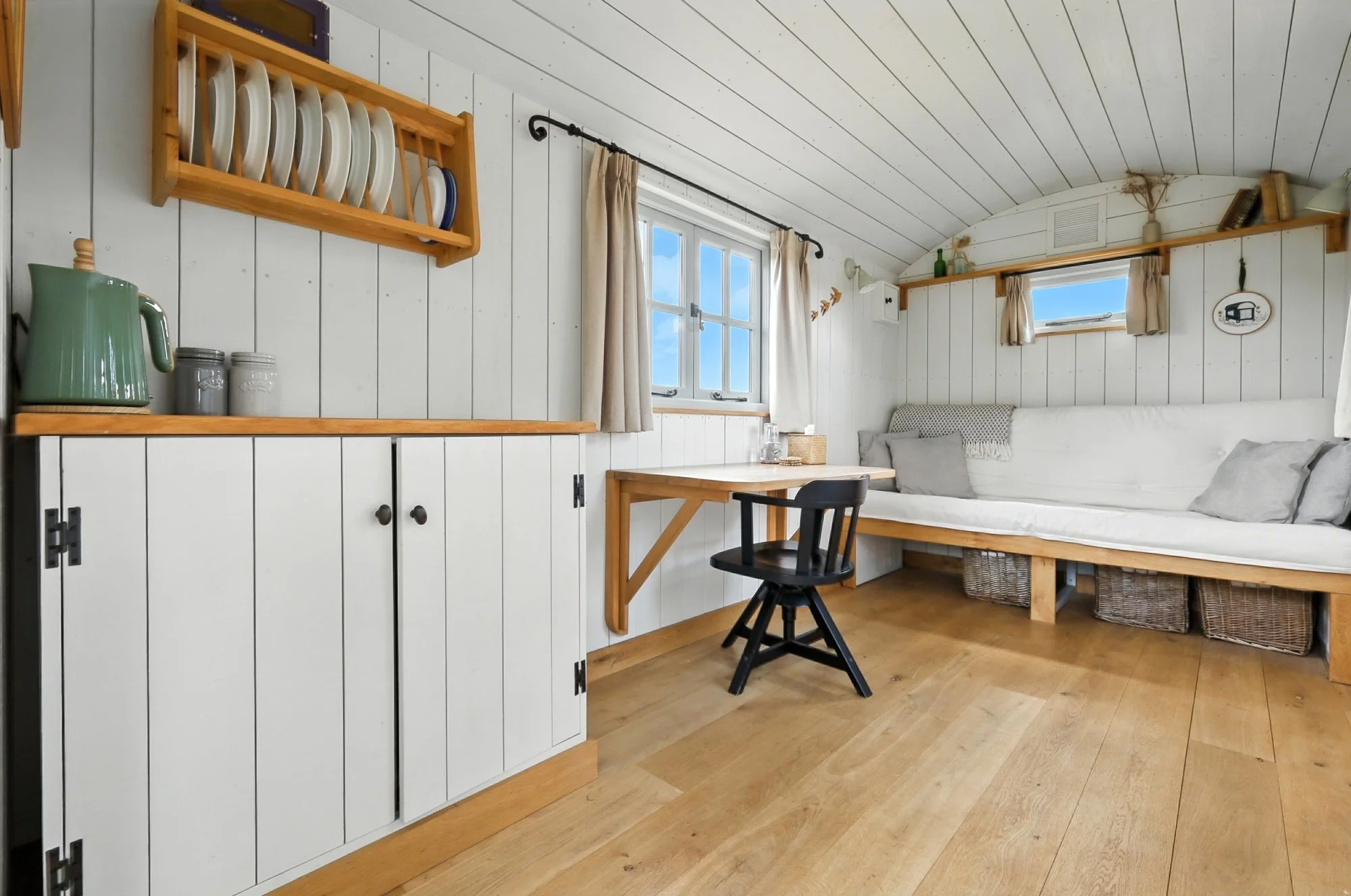 The image depicts a cozy, well-lit room with white wooden panel walls and ceiling. There is a window with beige curtains, a black chair at a wooden desk, and a white cushioned seating area along the wall. Wooden shelves hold decorative items and book